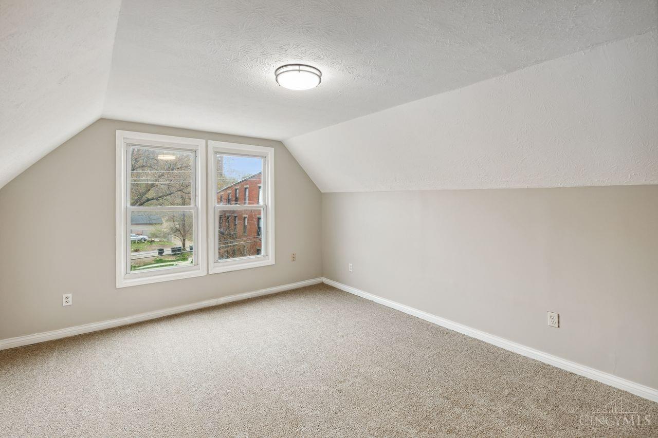 Vacant upstairs room with sloped ceilings, beige carpet, and two windows overlooking neighboring buildings.