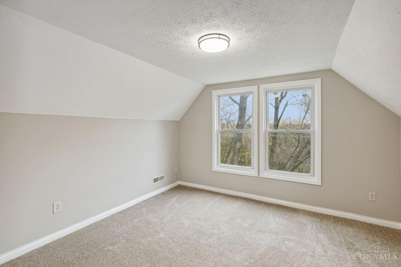 Vacant carpeted room with a sloped ceiling and two windows overlooking trees.
