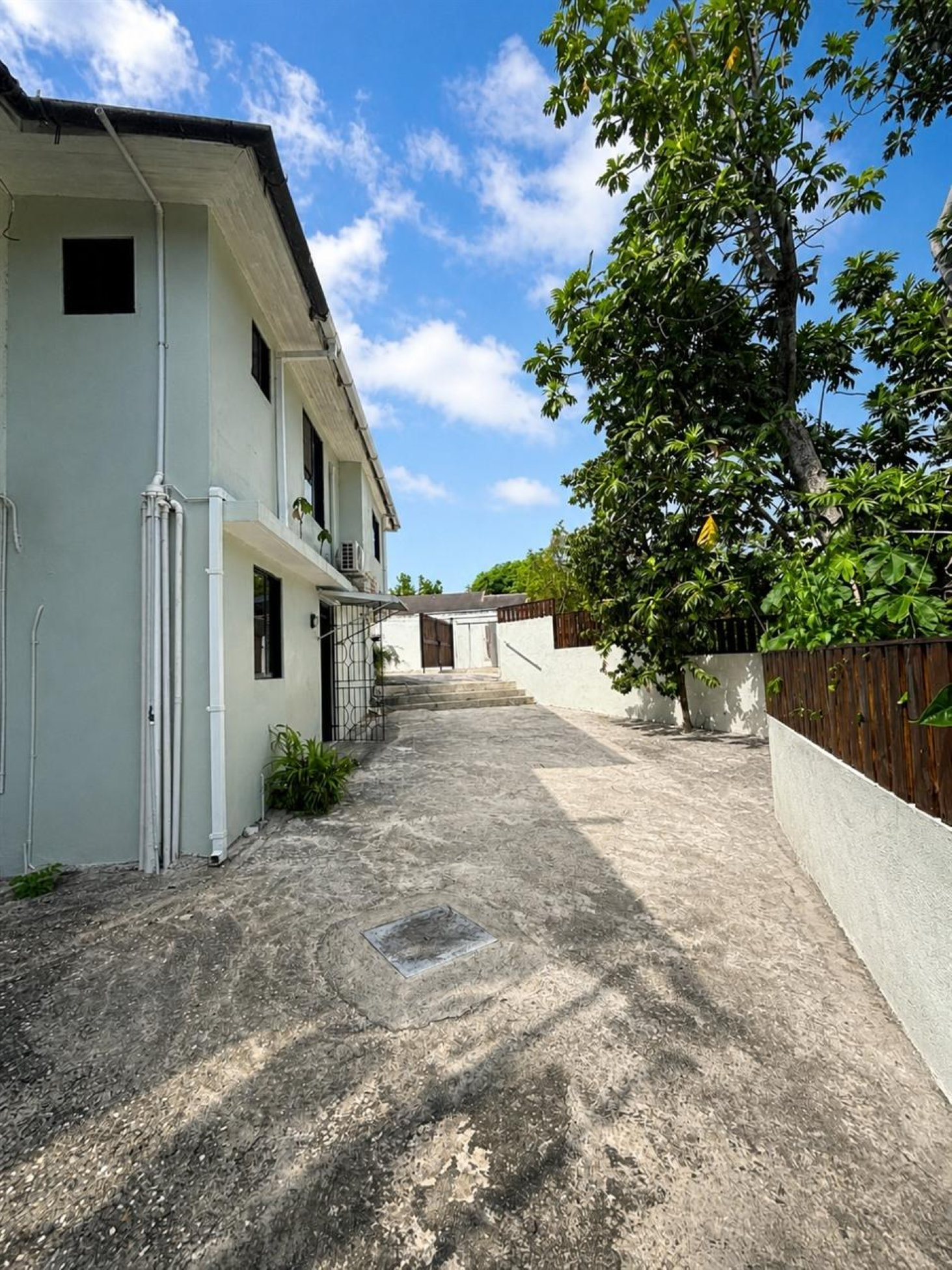 Two-story house beside a paved driveway bordered by leafy trees and wooden fencing.