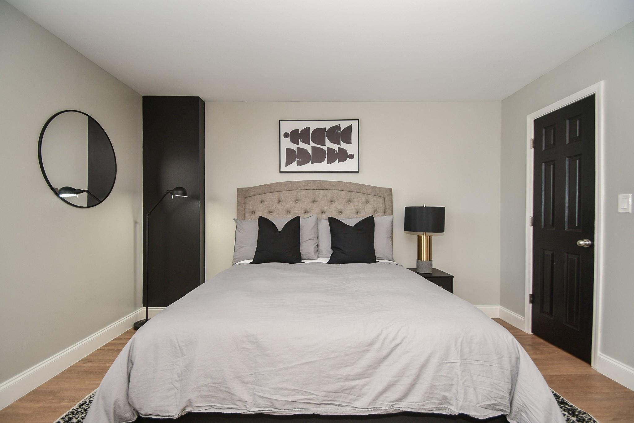 Staged bedroom with a gray bed, tufted headboard, black accent pillows, and round wall mirror.