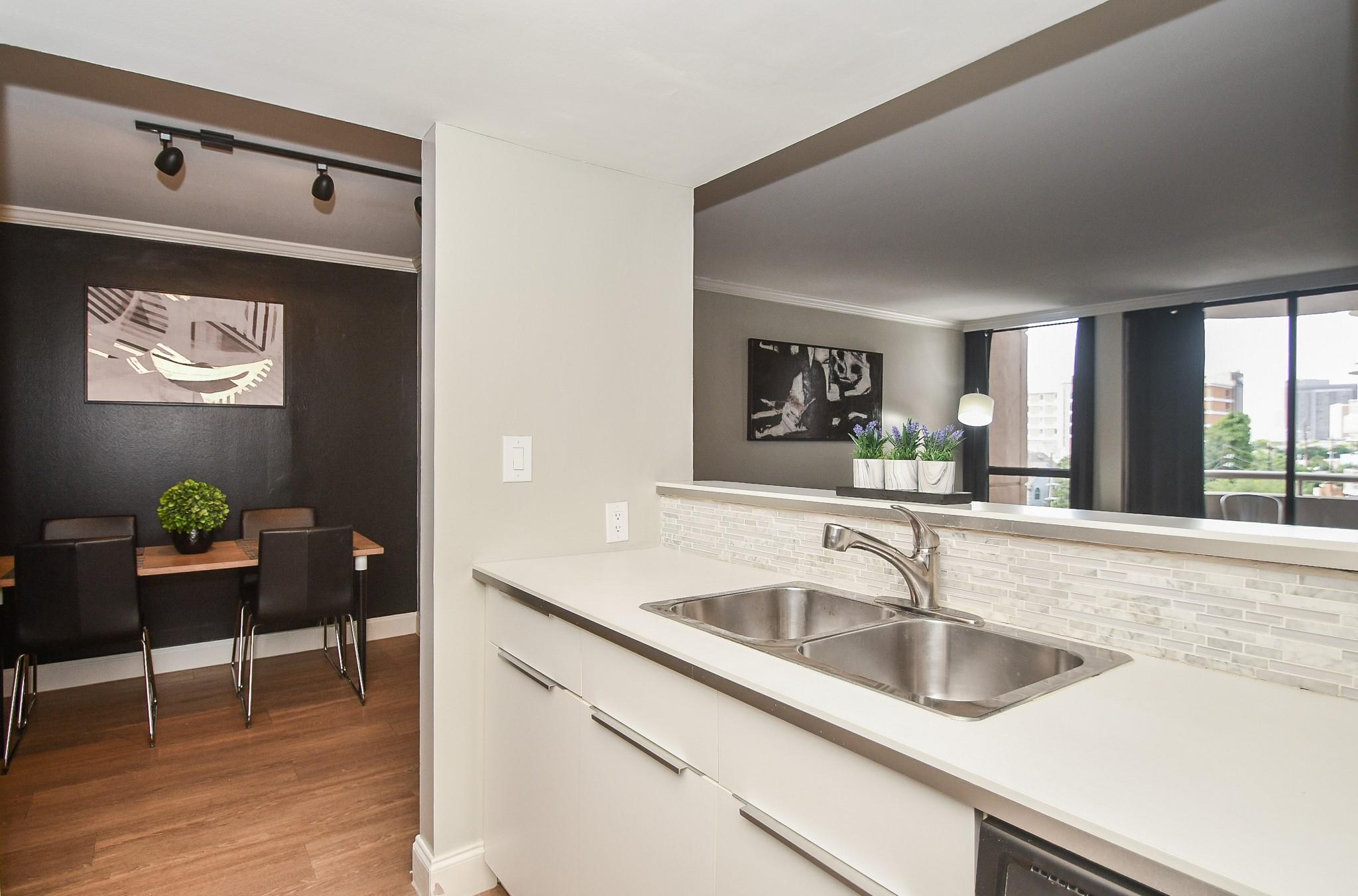 Condo kitchen with a double-basin sink and light backsplash beside a dining area with dark chairs.