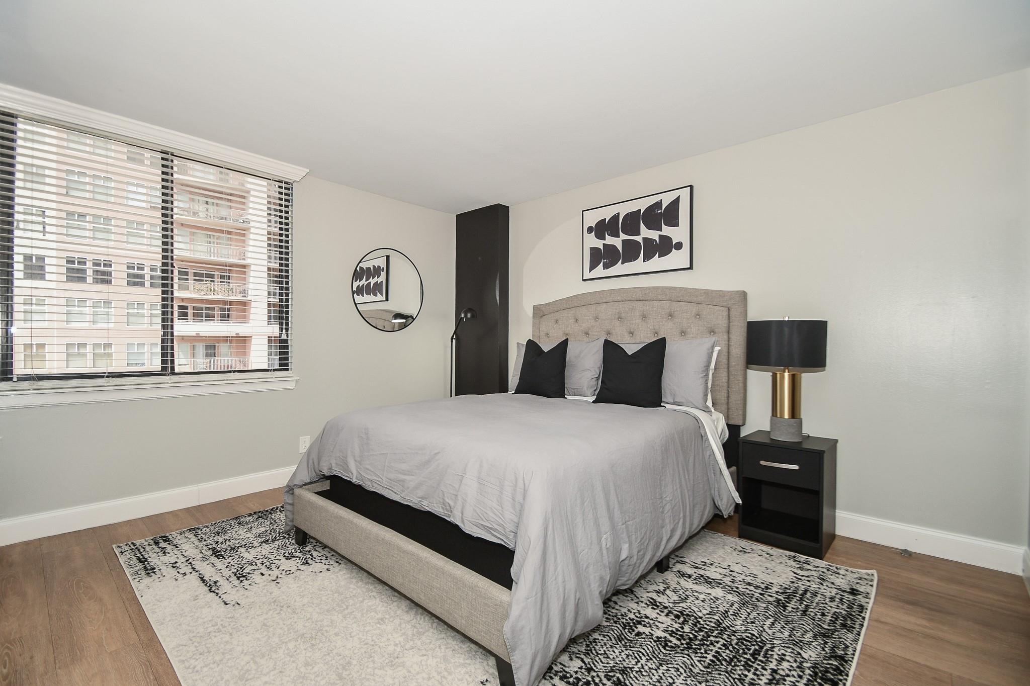 Bedroom with a gray upholstered bed, patterned rug, and large window with blinds.