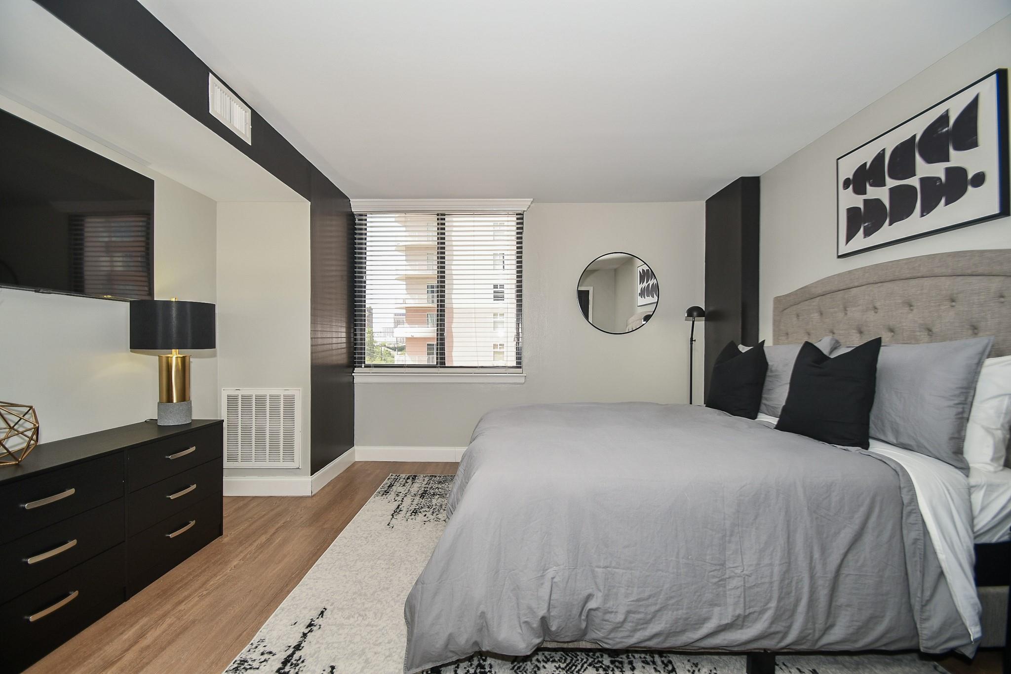 Furnished bedroom with gray bedding, black accent pillows, and a dark dresser beside the window.