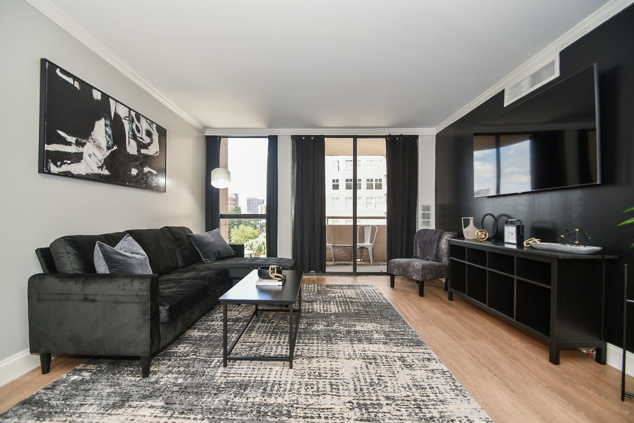 Furnished living room with a dark sectional, large patterned rug, and windows overlooking nearby buildings.