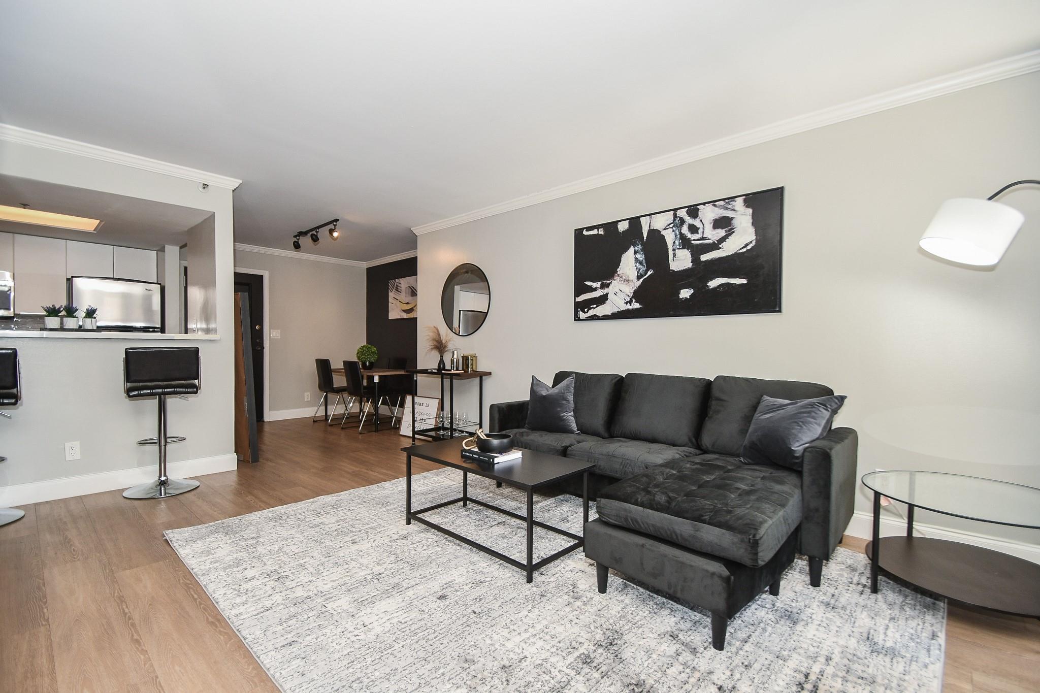 Furnished living room with a dark sectional, light rug, and open dining and kitchen areas.