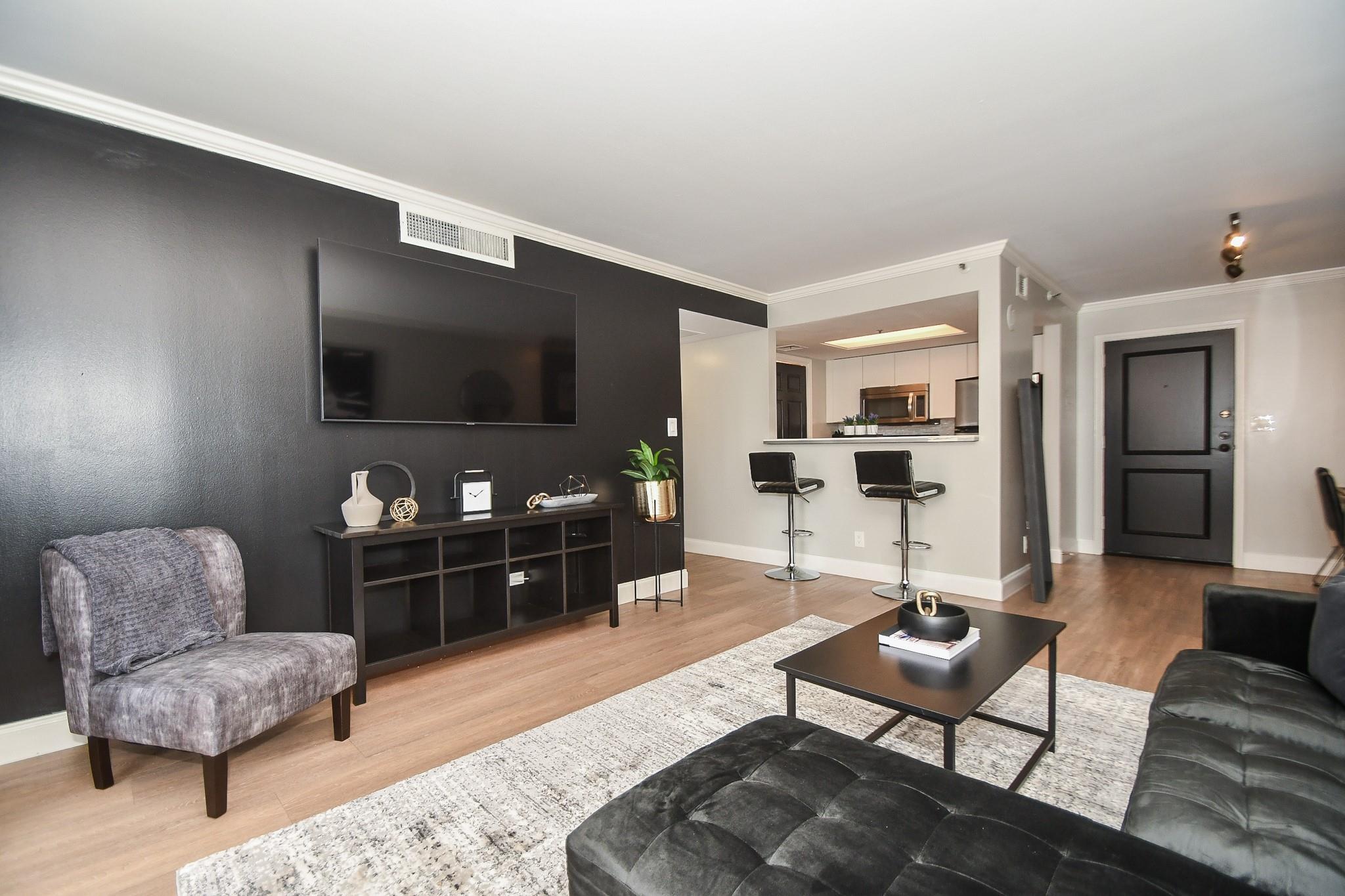 Staged condo living room with charcoal accent wall, wall-mounted television, and open kitchen breakfast bar.