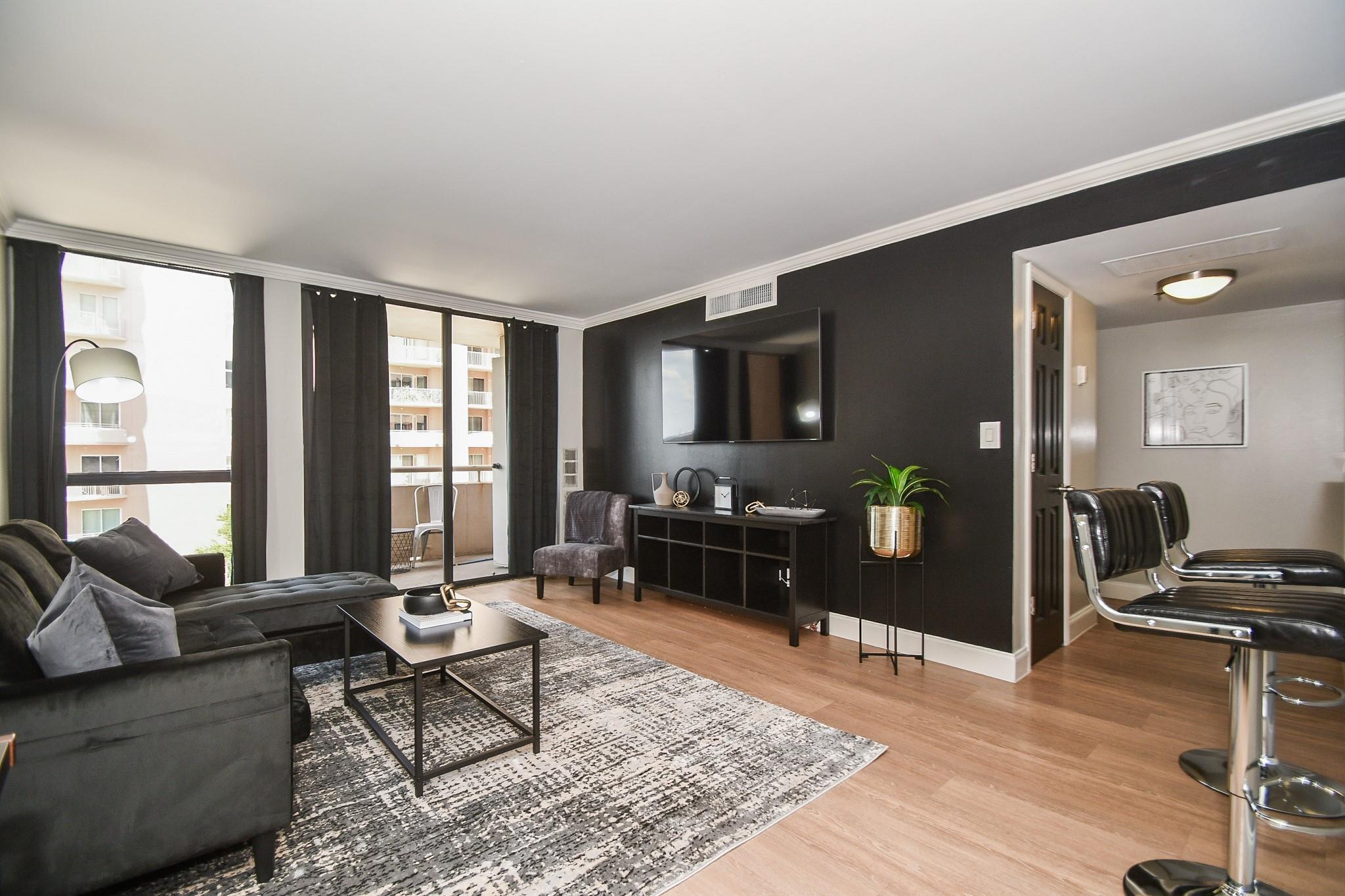 Staged condo living room with charcoal sofa, wall-mounted TV, and windows opening to a balcony.