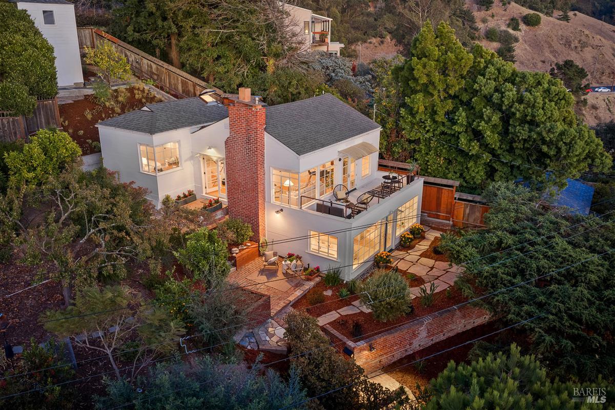 White two-story home with a brick chimney, glowing windows, and a furnished patio at dusk.