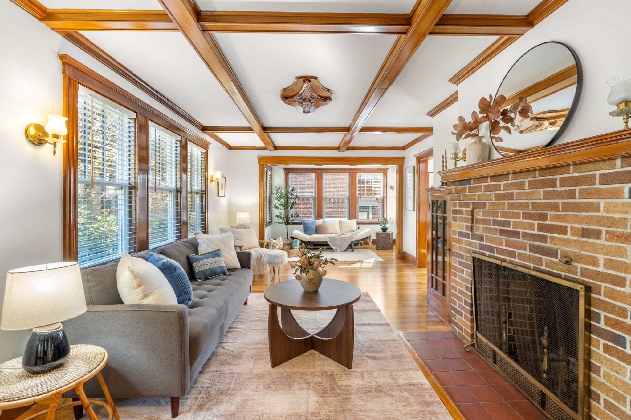 Staged living room with a brick fireplace, wood-beamed ceiling, and large windows.