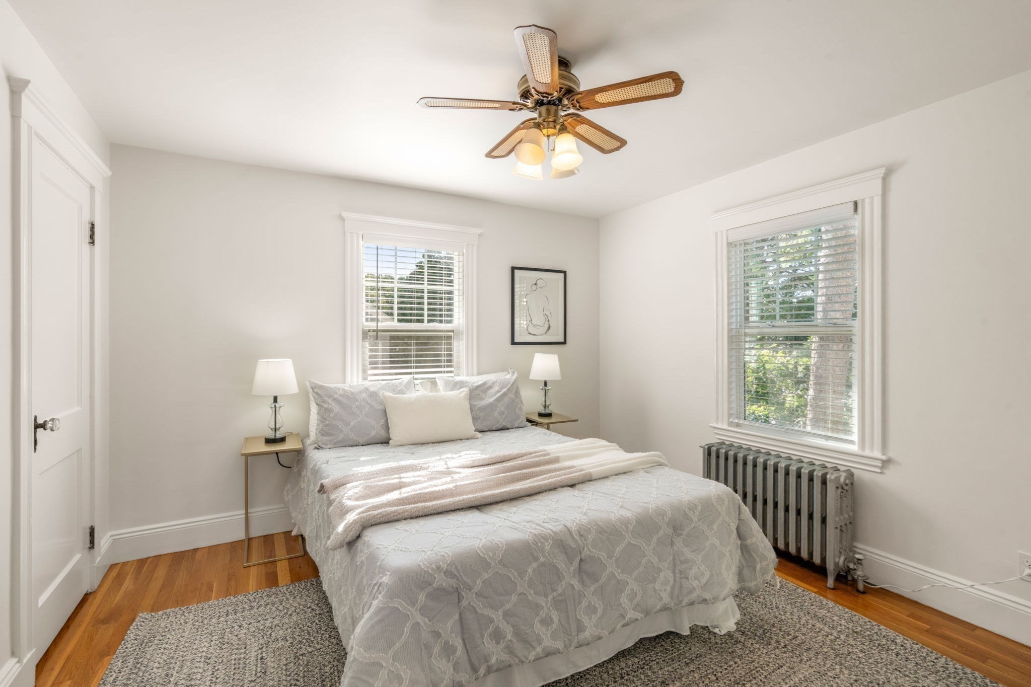 Staged bedroom with a patterned bed, two bright windows, hardwood floors, and a cast-iron radiator.