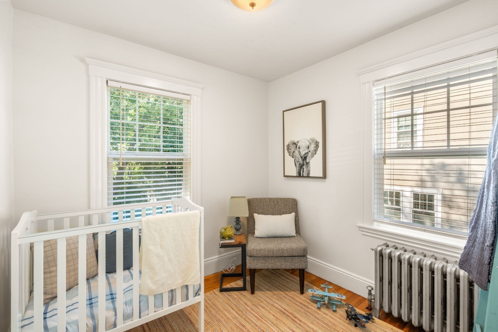 Nursery with a white crib, upholstered chair, elephant artwork, and two sunlit windows.