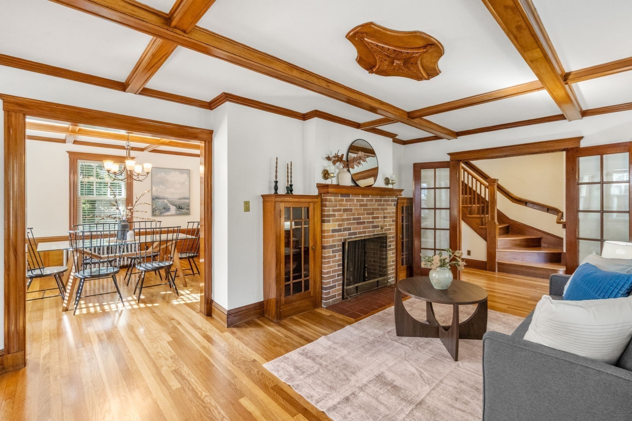 Living room with a brick fireplace, wood-beamed ceiling, and staircase beside the dining room.