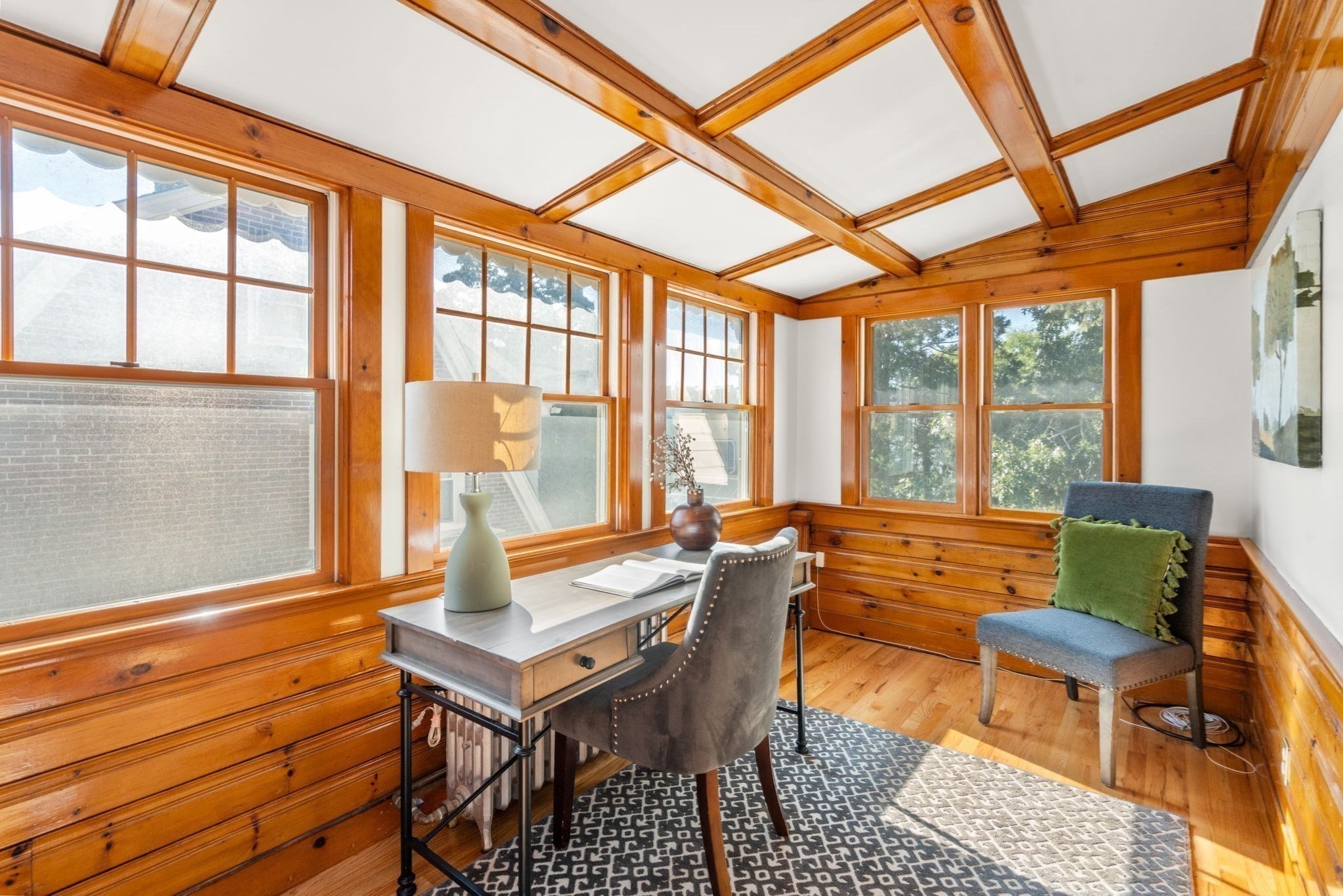 Sunroom with warm wood paneling, timber ceiling beams, large windows, and a desk with chairs.