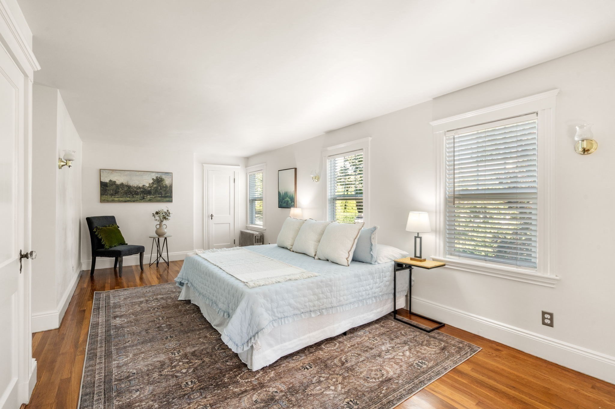 Bright bedroom with pale blue bedding, patterned rug, wood floors, and windows with blinds.