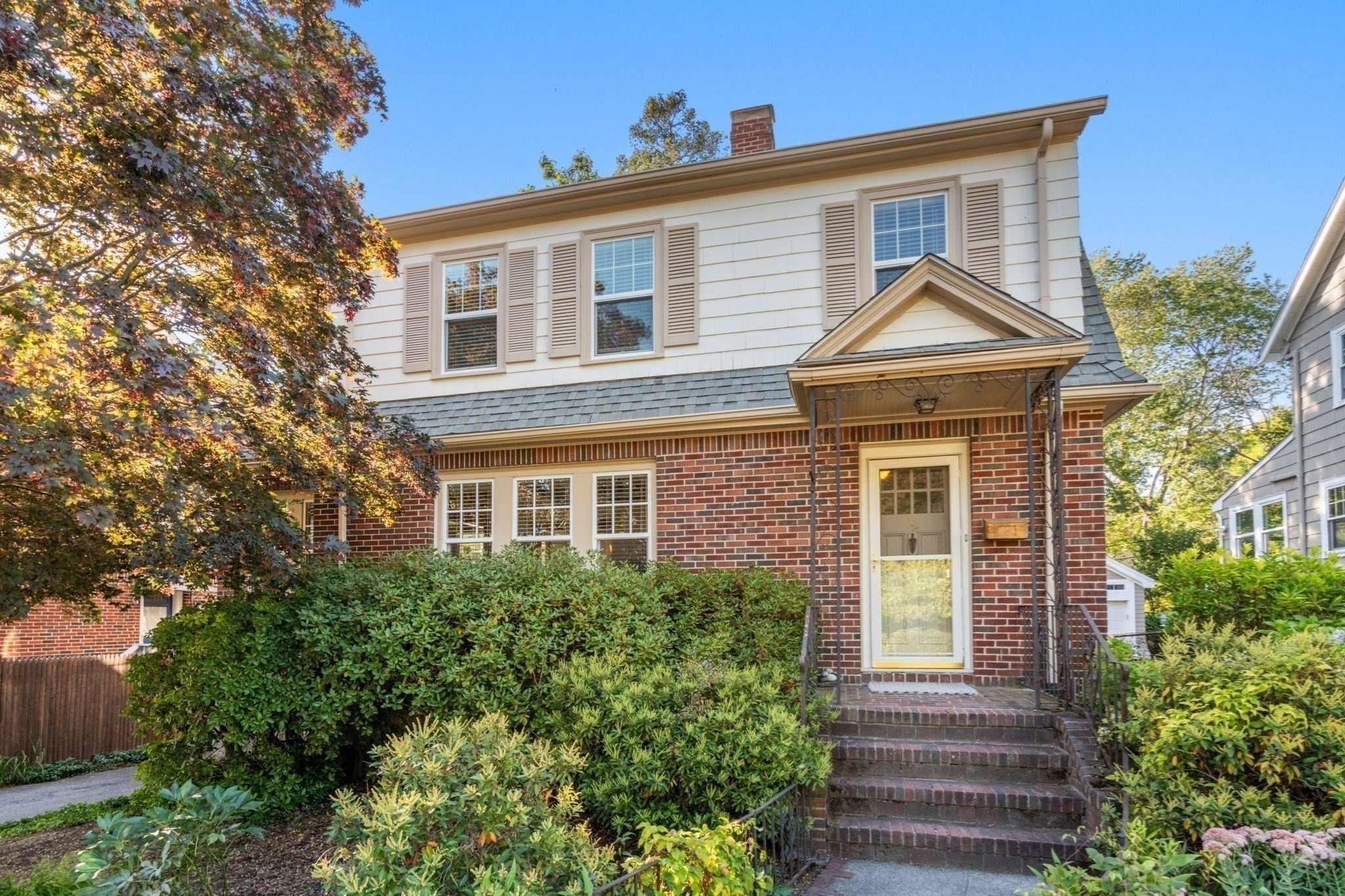 Two-story brick and white-shingle house with a covered entry and brick steps amid leafy landscaping.