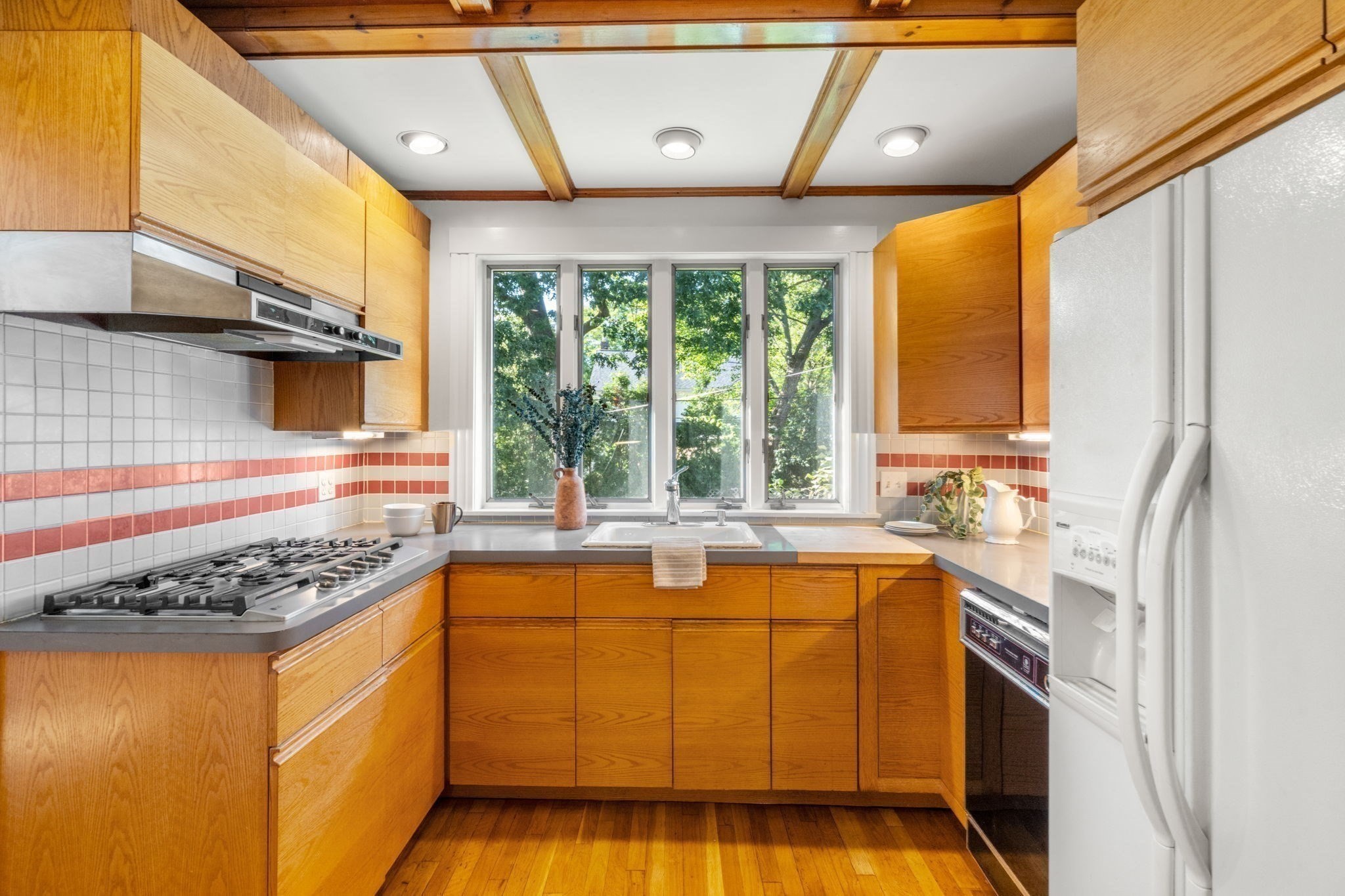 Kitchen with honey-colored wood cabinets, red-striped tile backsplash, and a sink beneath a tree-lined window.
