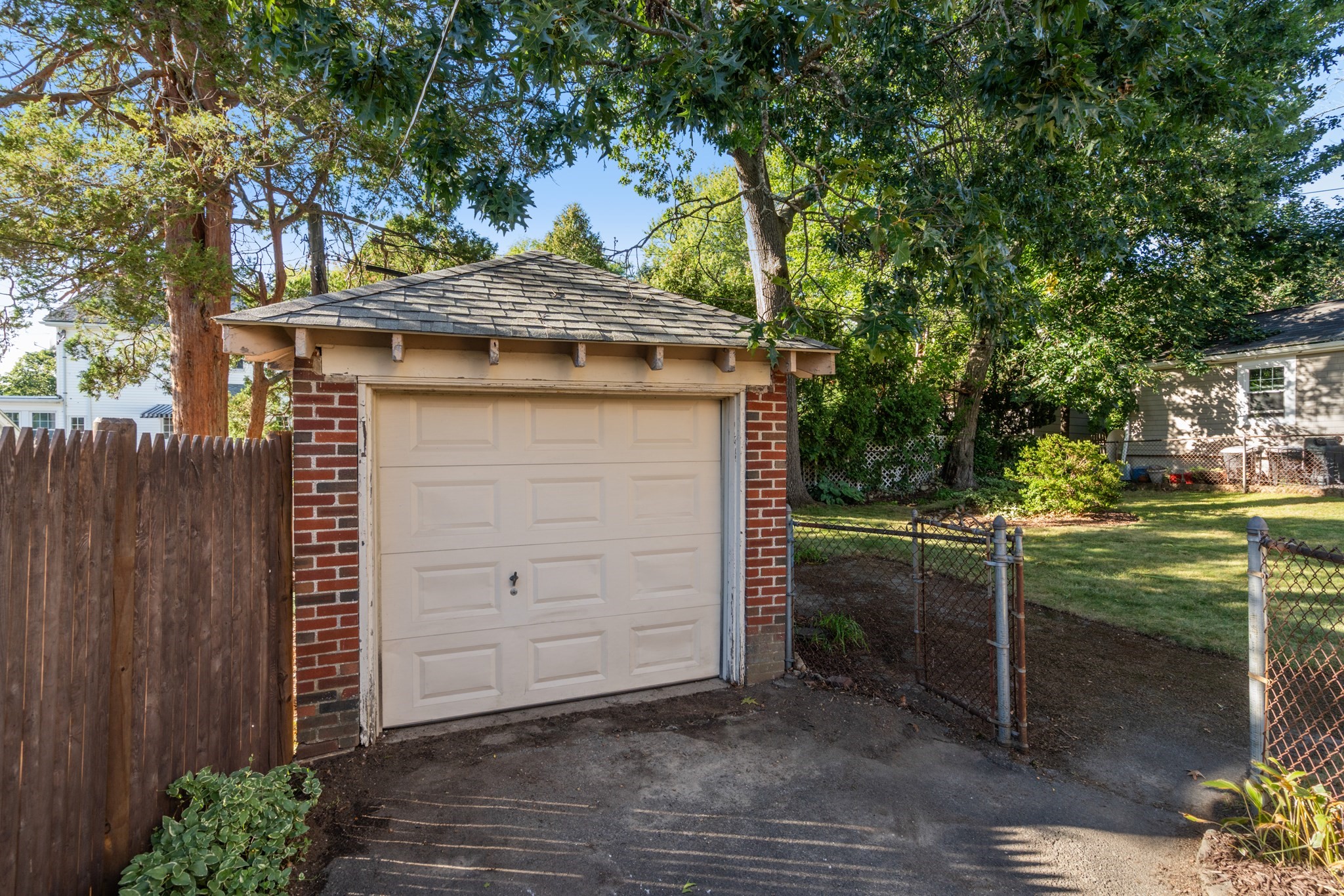 Detached brick garage with a beige overhead door beside a fenced, tree-shaded yard.