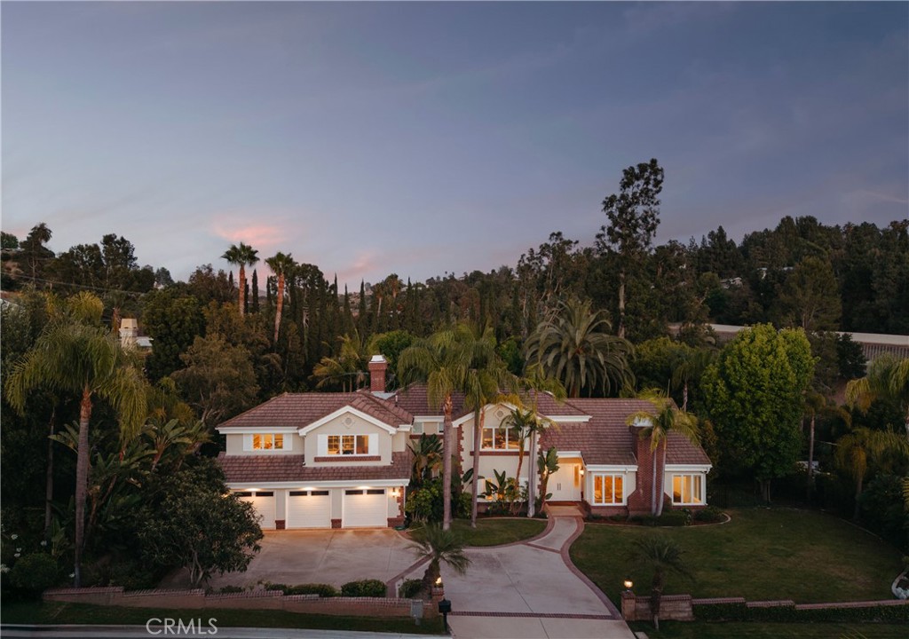 Two-story home with a tiled roof, three-car garage, curved driveway, and palm-lined landscaping at dusk.