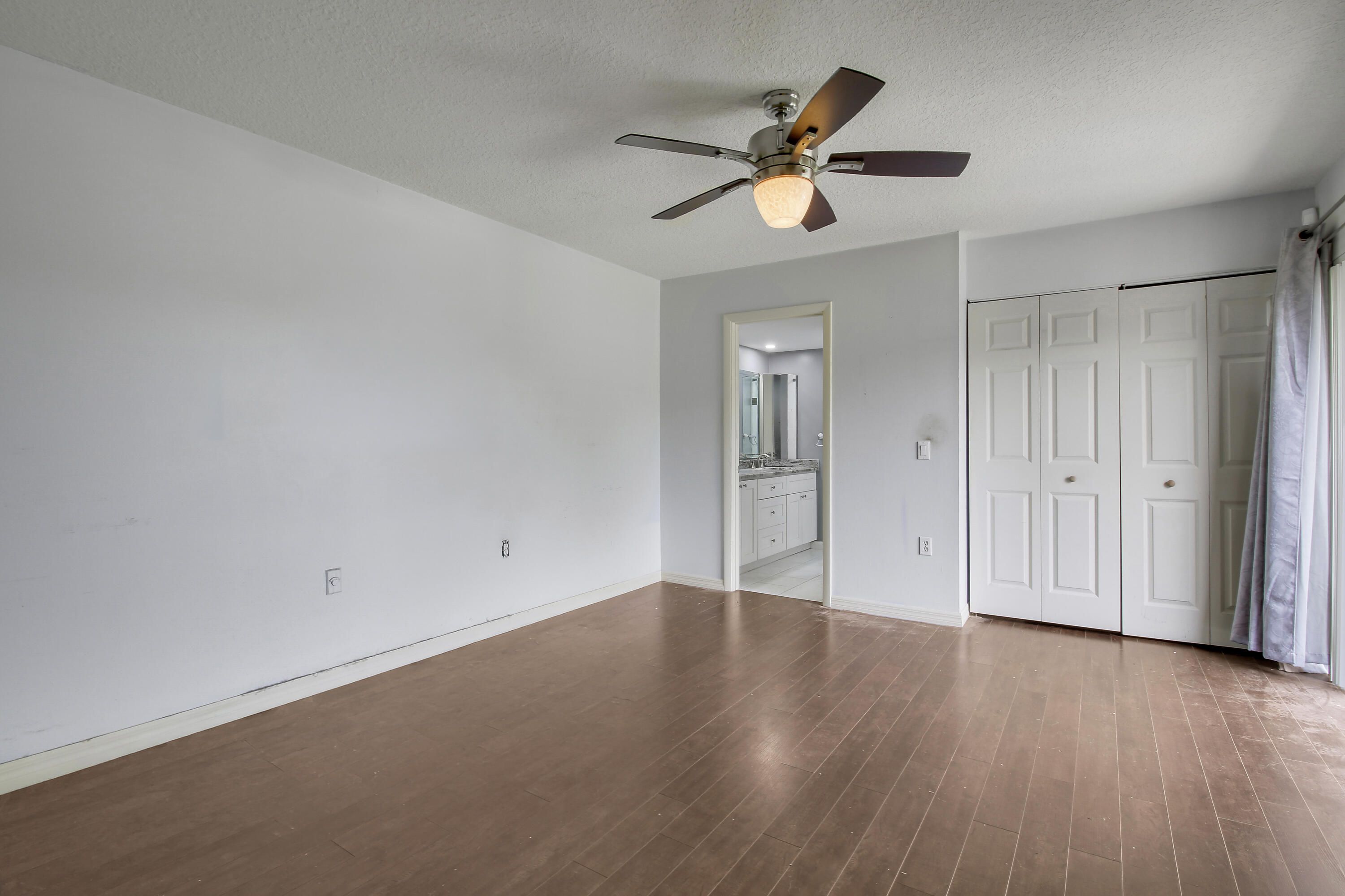 Empty bedroom with brown plank flooring, ceiling fan, large closet, and doorway to an en-suite bathroom.