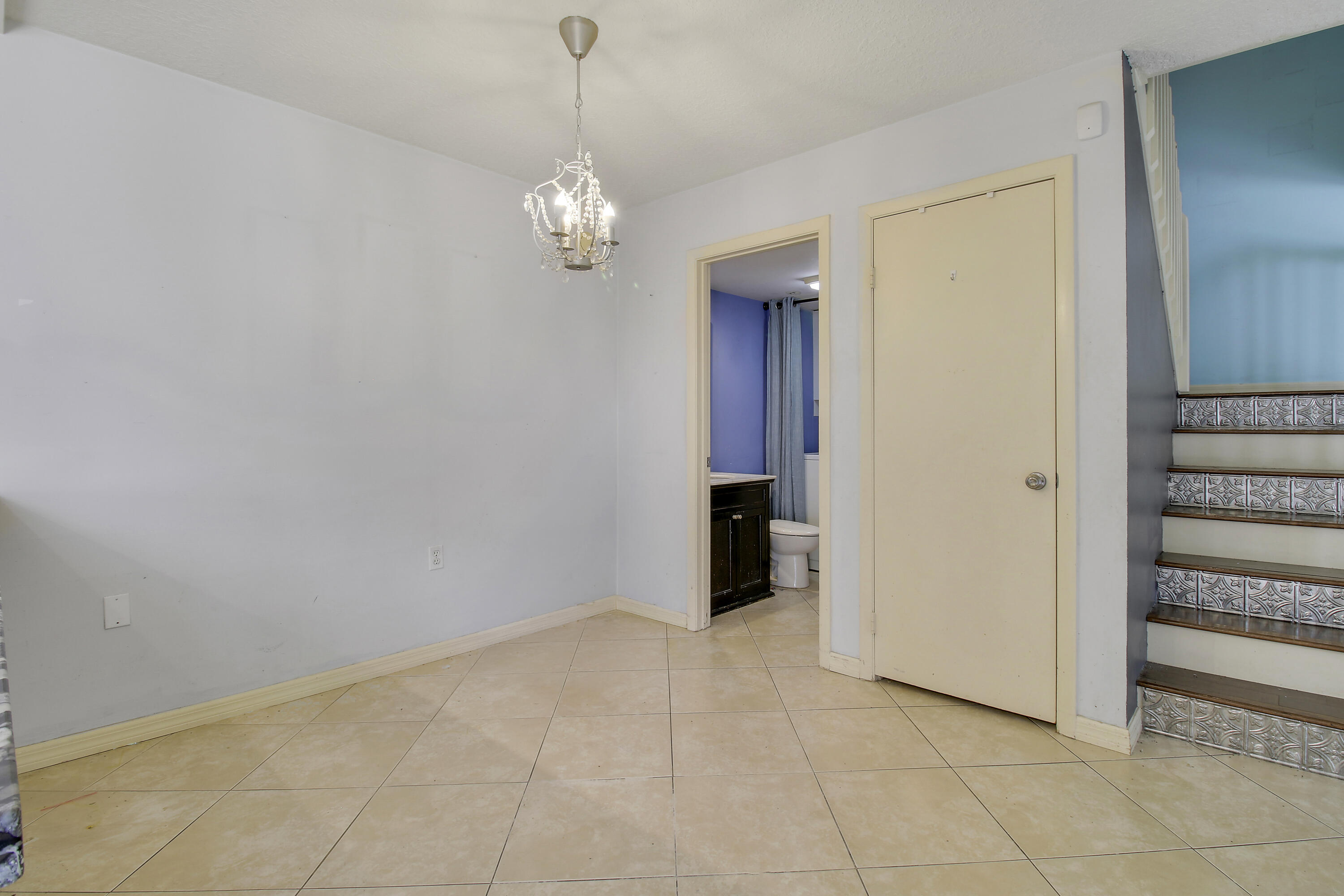 Empty tiled room with a chandelier, bathroom doorway, and staircase with patterned risers.