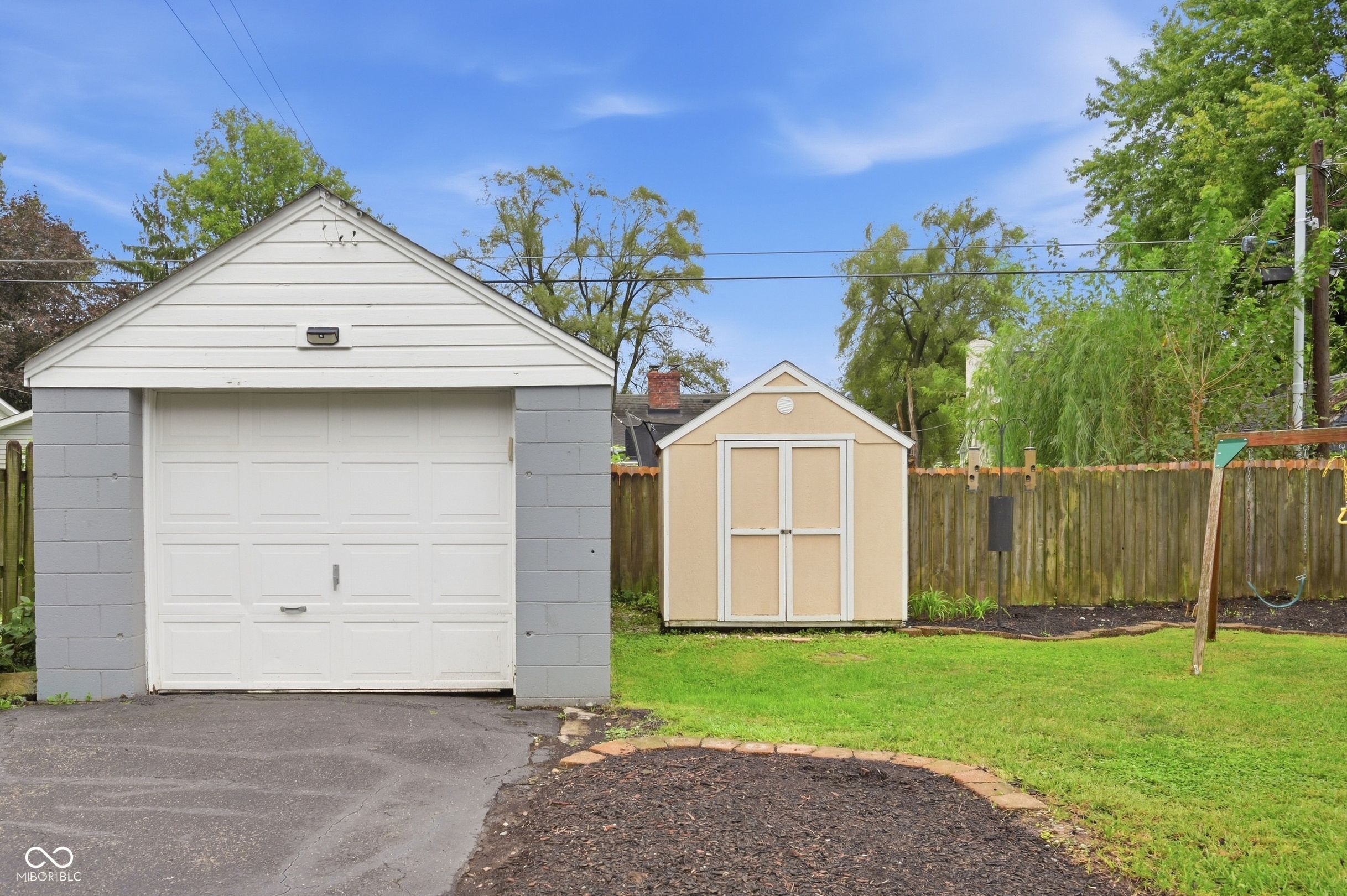 Backyard with a white detached garage, tan storage shed, wooden fence, and swing set.