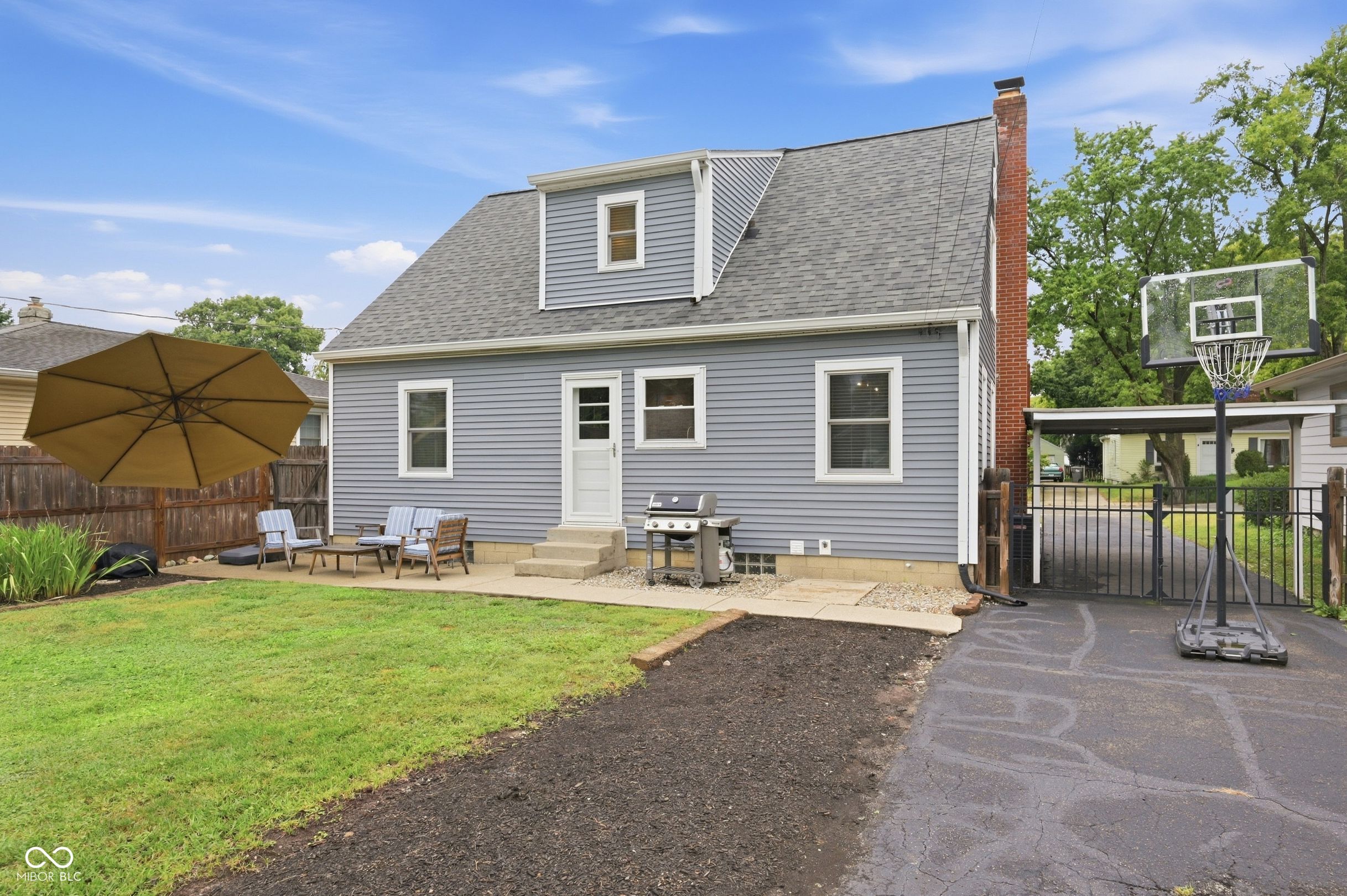 Gray house’s rear exterior with a patio, grill, umbrella, and basketball hoop beside the driveway.