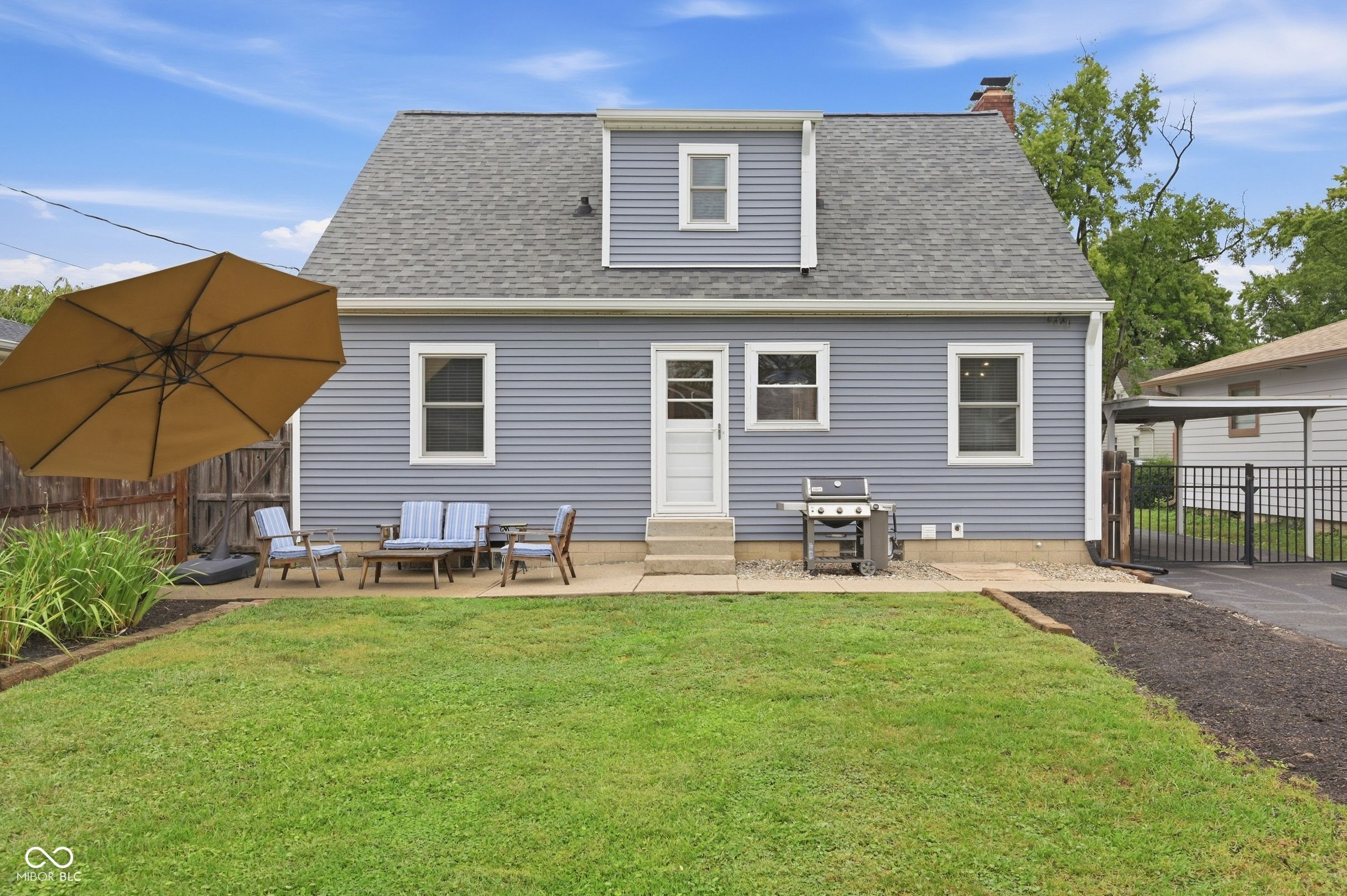 Gray house rear exterior with patio seating, umbrella, barbecue grill, and green lawn.