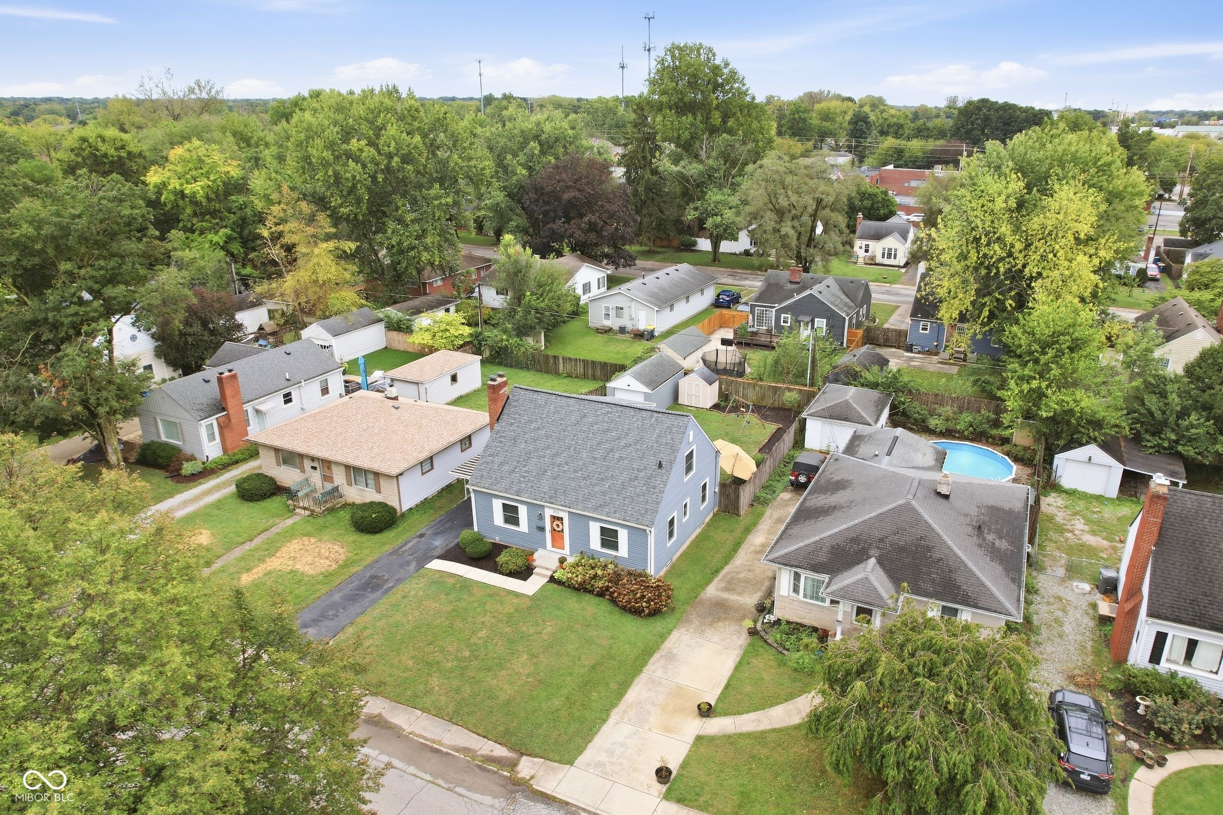 Aerial view of a blue-sided house among tree-lined Indianapolis homes, with fenced yards and a nearby pool.
