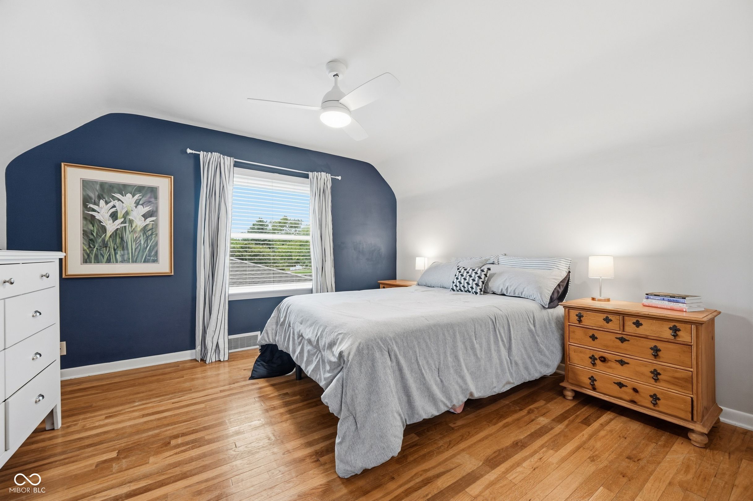 Bedroom with a gray bed, navy accent wall, hardwood floors, and ceiling fan.