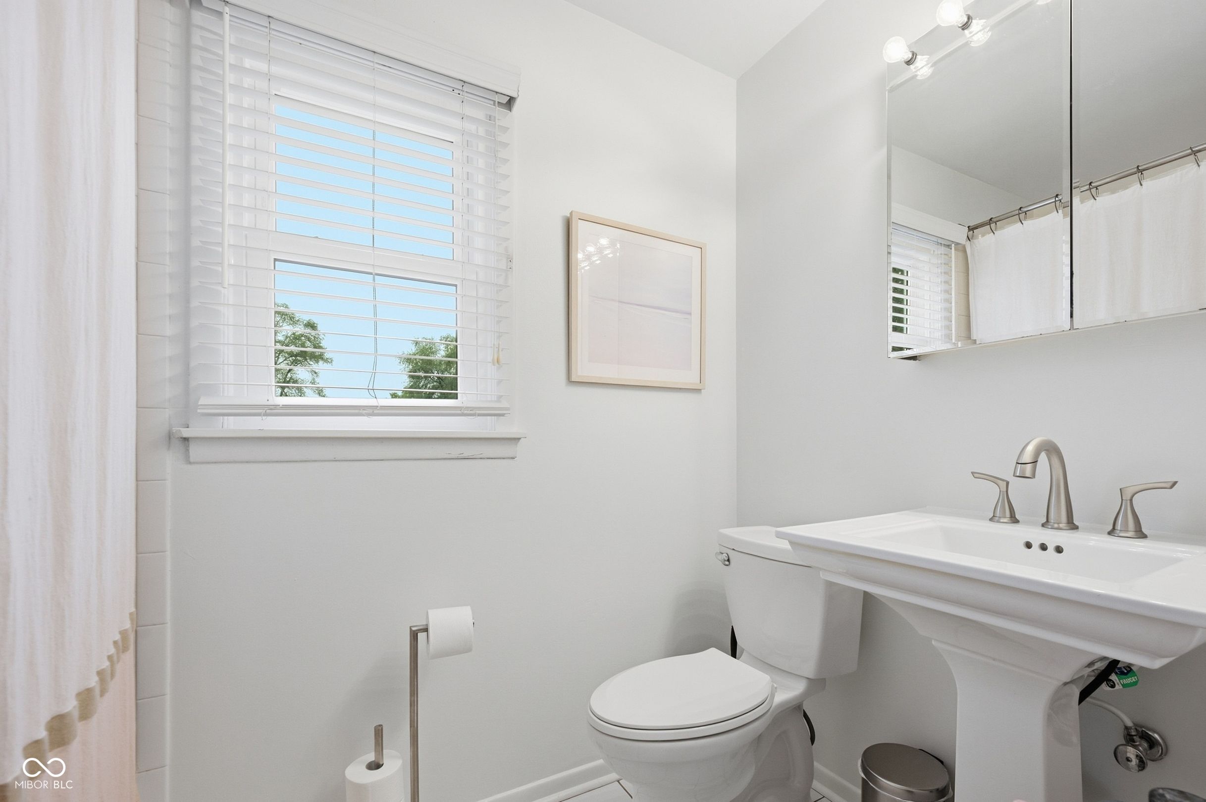 Bright bathroom with a pedestal sink and toilet beneath a large mirror and window with white blinds.