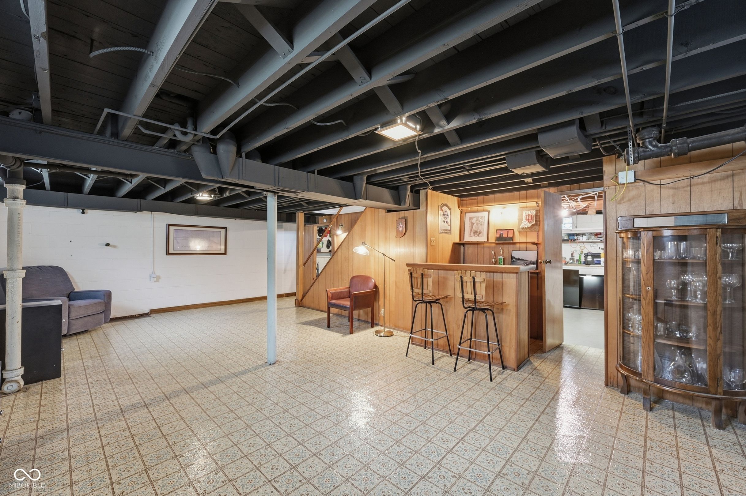 Basement recreation area with a wood-paneled bar, two stools, tiled floor, and exposed ceiling beams.