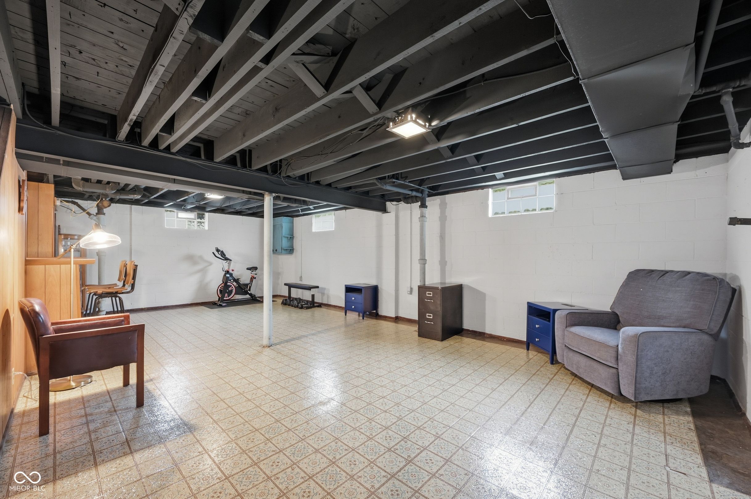 Furnished basement with exposed beams, patterned tile flooring, an exercise bike, and a gray armchair.