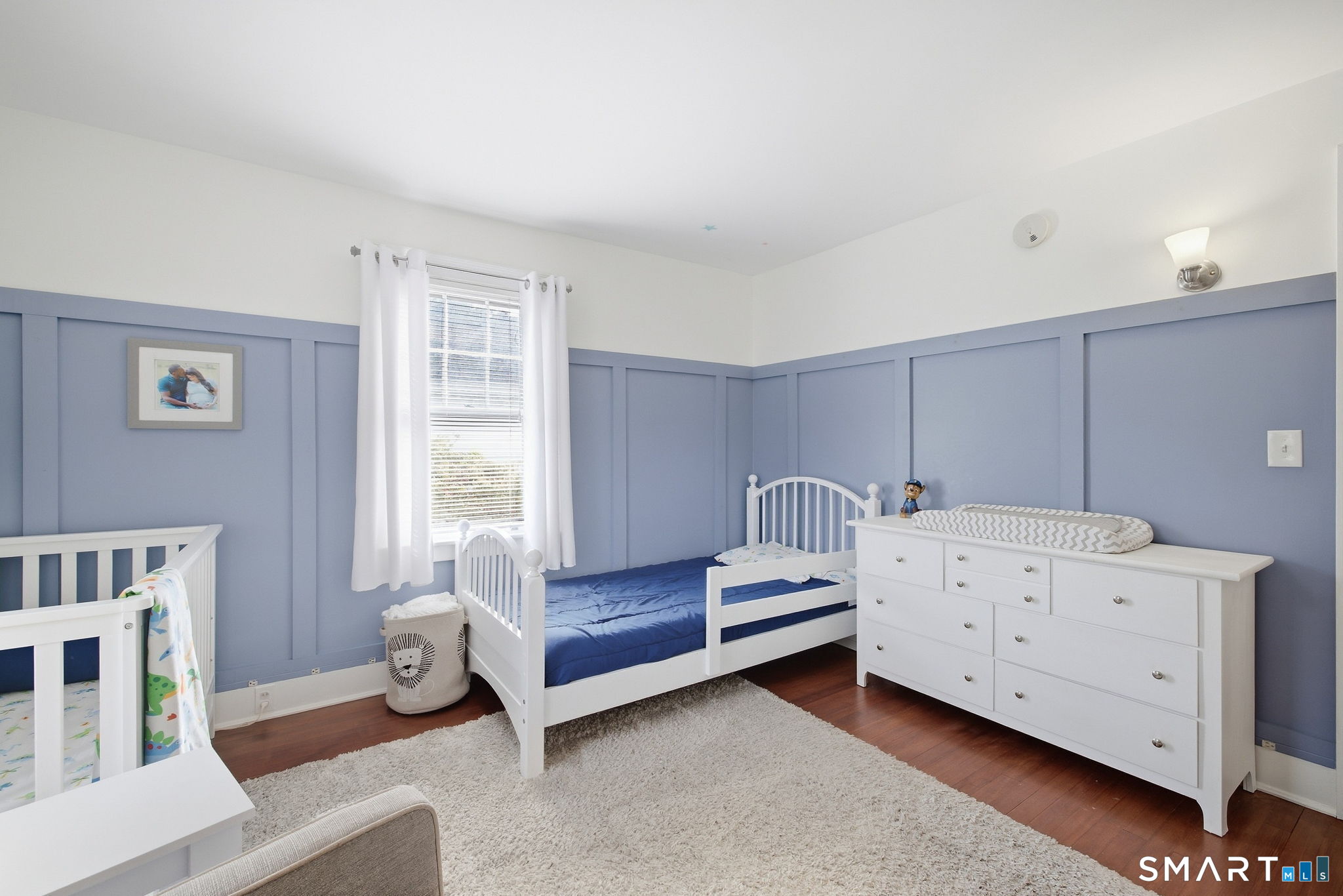 Nursery with blue paneled walls, white crib and bed, and a large white dresser.