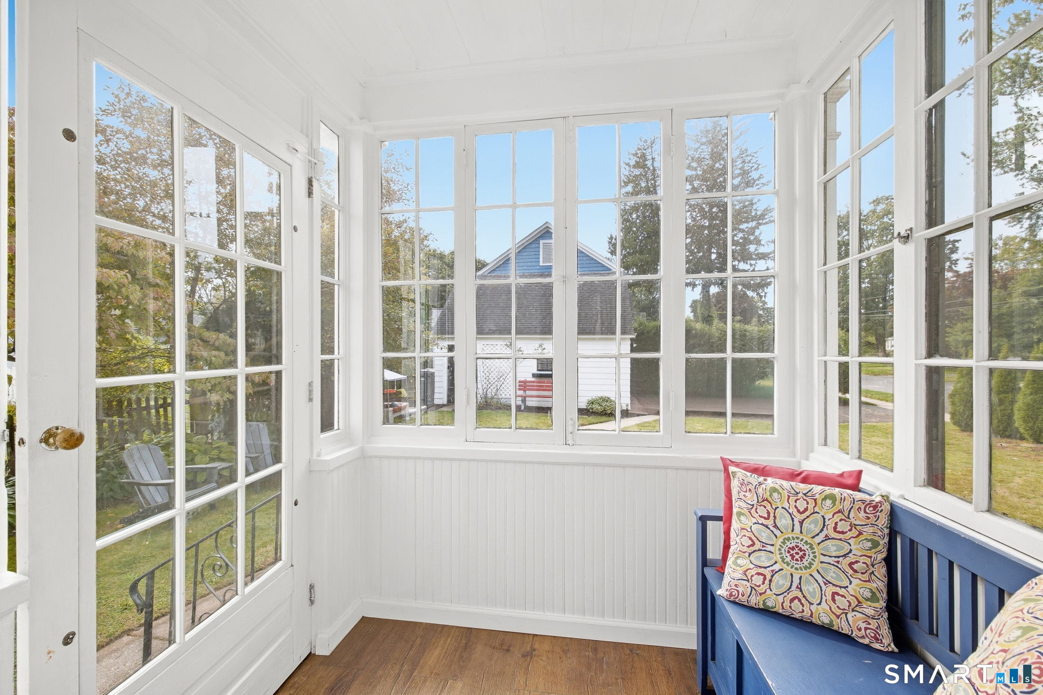 Enclosed porch with bright white grid windows, wood flooring, and a blue bench with colorful cushions.