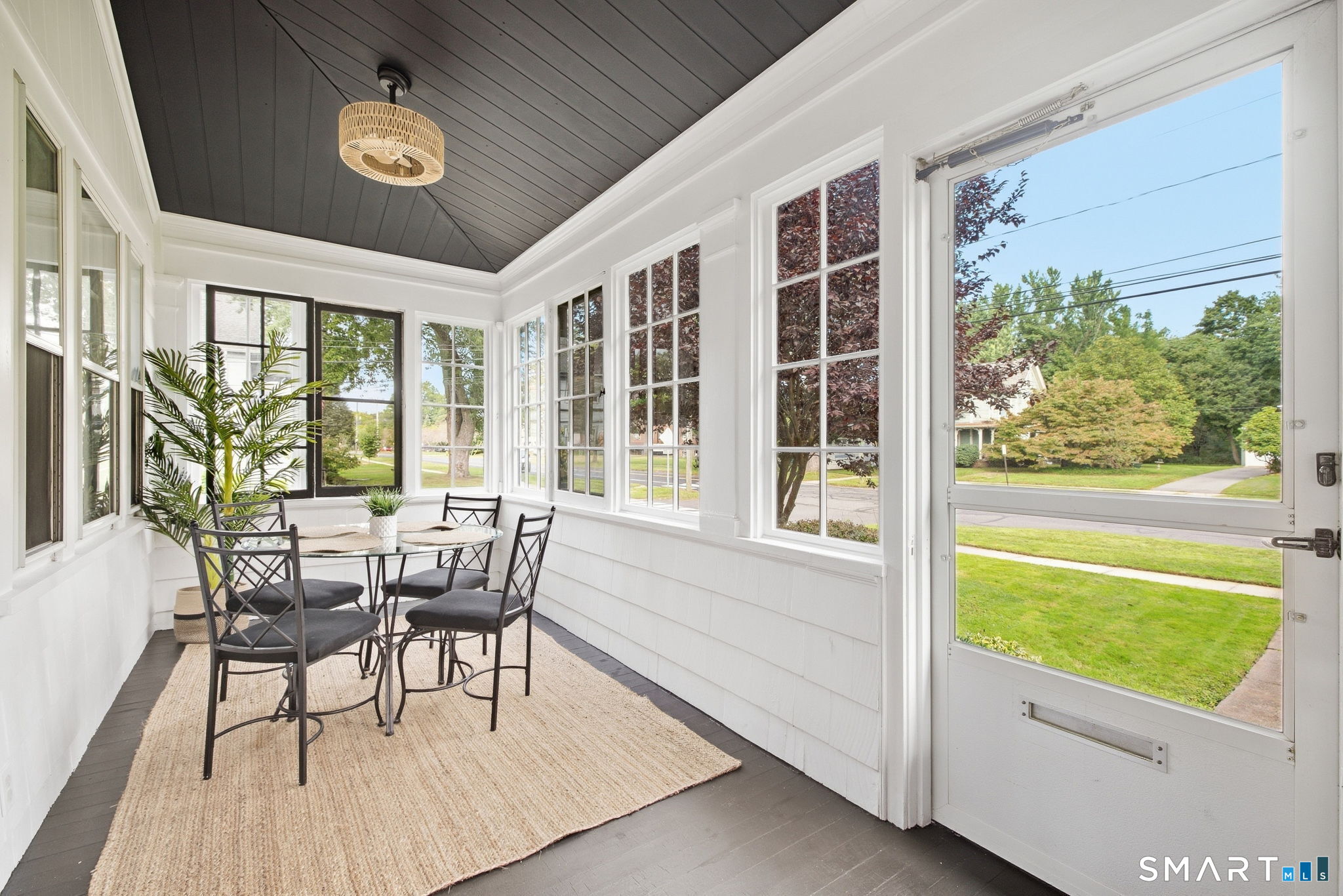 Furnished enclosed porch with a black dining set, woven rug, and windows overlooking a green lawn.