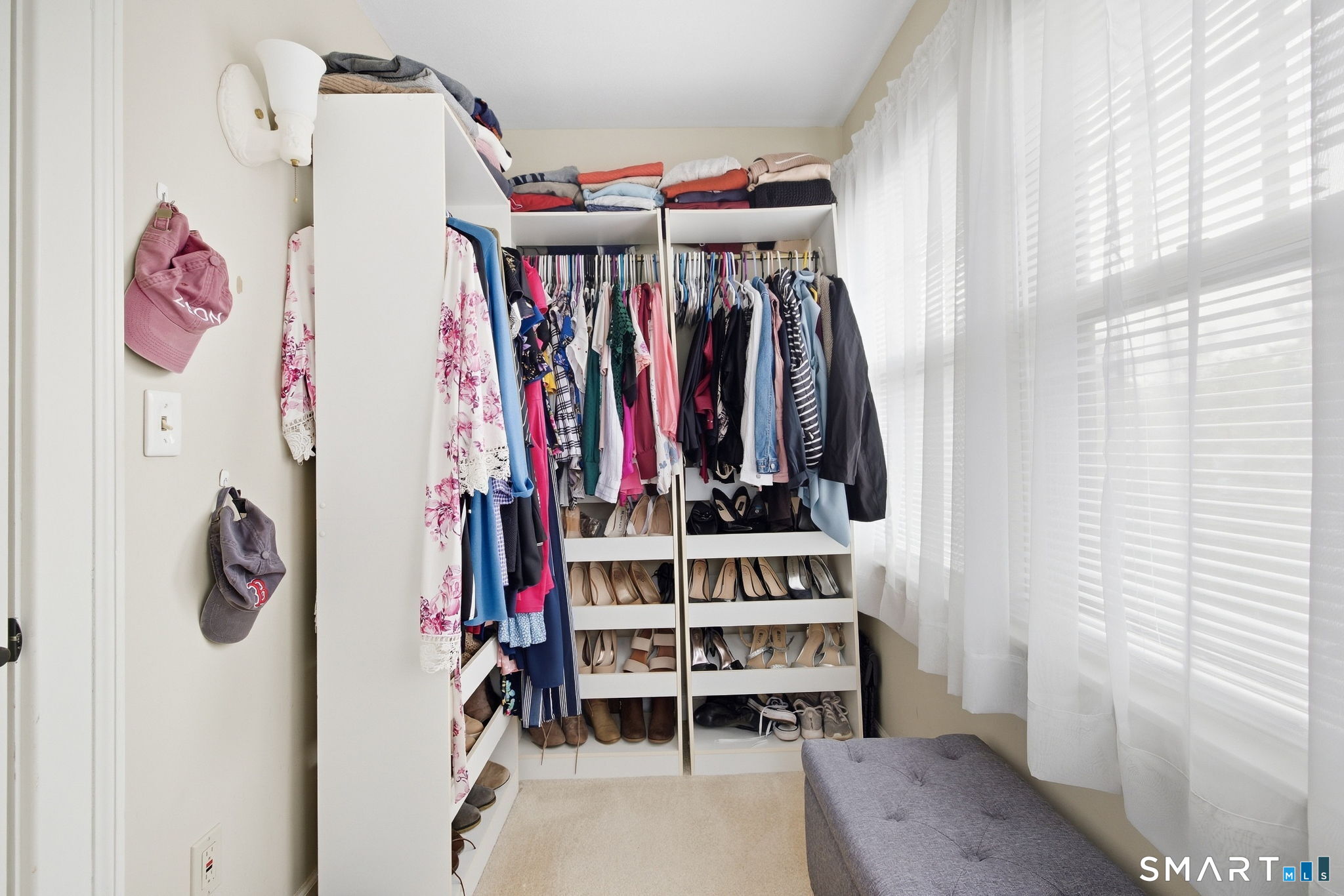 Walk-in closet with white clothing and shoe shelves, bright curtained windows, and a gray upholstered bench.
