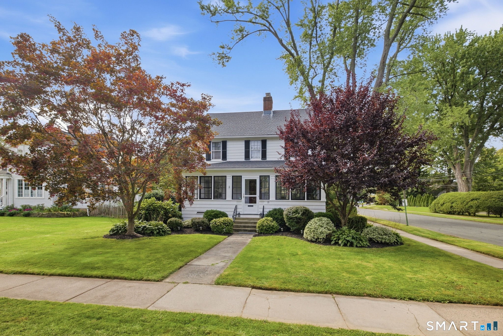 White two-story house with black shutters, a covered porch, and mature trees framing the landscaped front lawn.