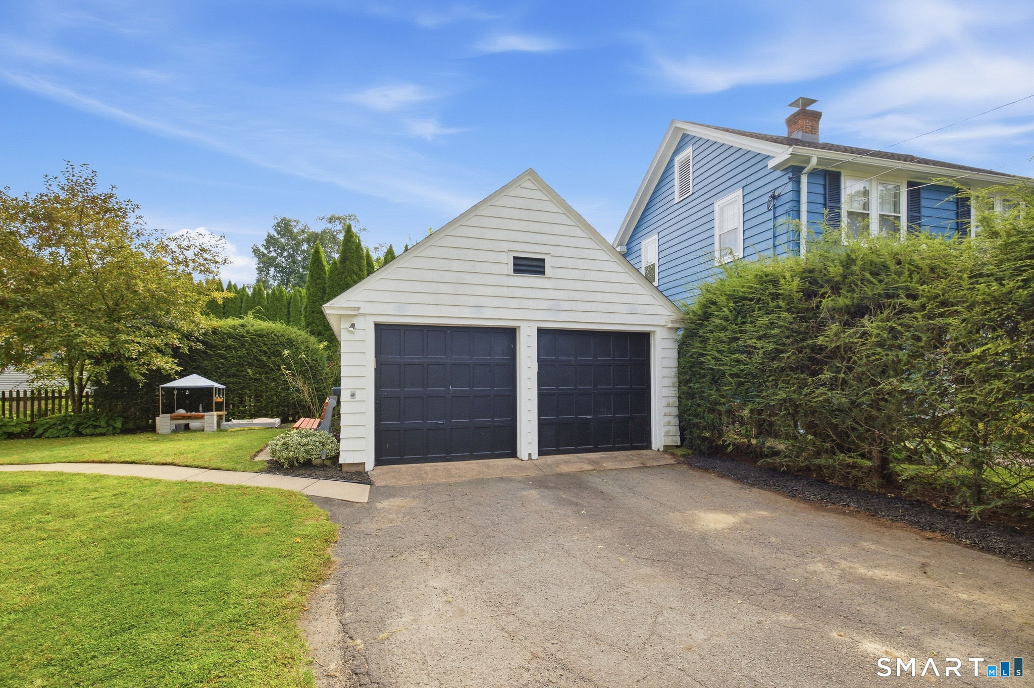 White two-car garage with dark doors beside a blue-sided house and hedge-lined driveway.