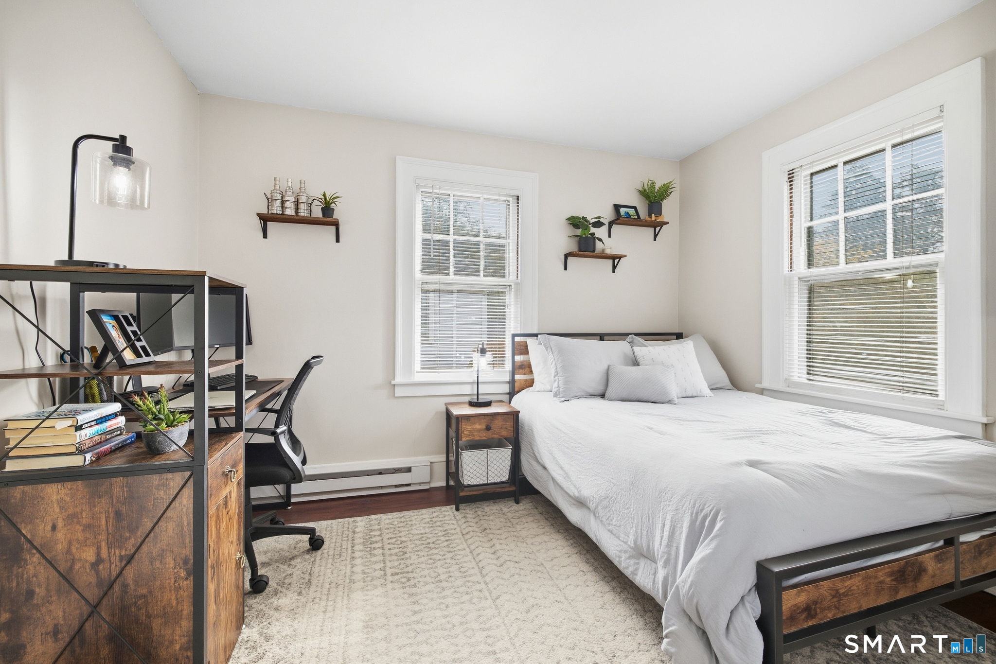 Furnished bedroom with a white bed, two bright windows, and a wood-and-metal desk.