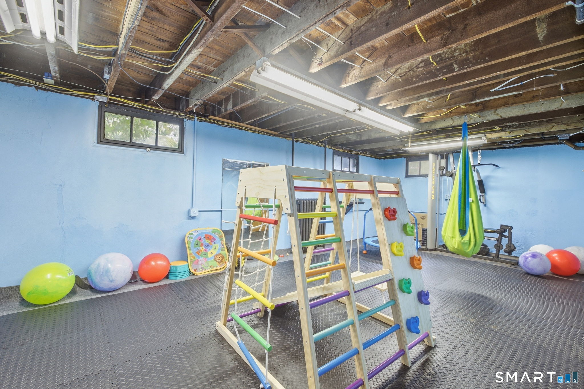 Basement playroom with a colorful wooden climbing structure, exercise balls, and a hanging fabric swing.