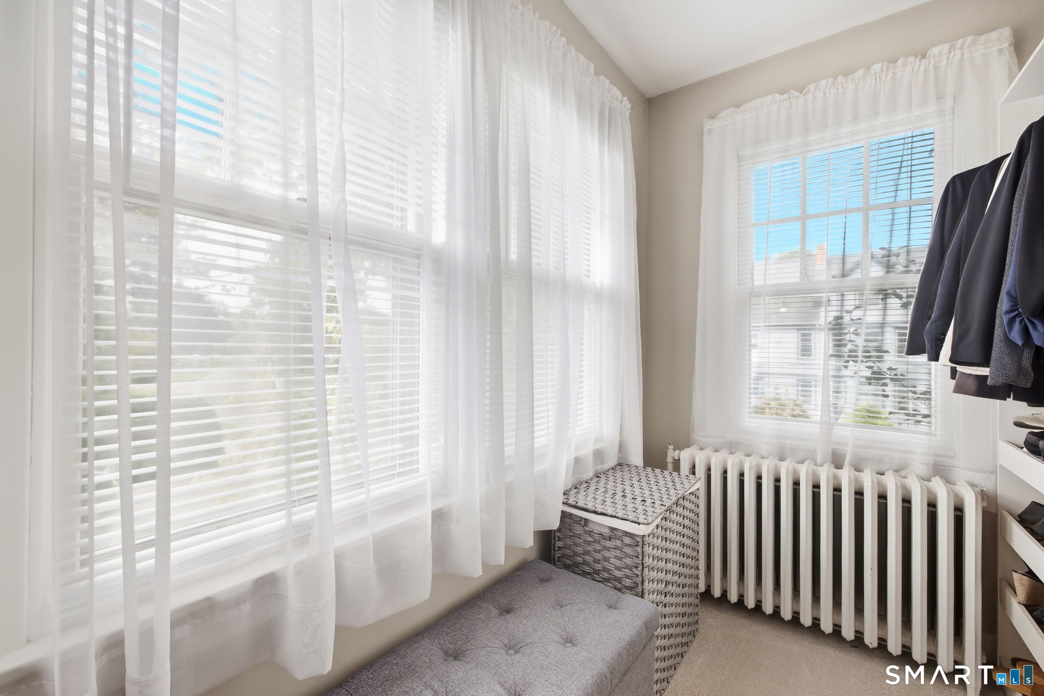 Bright dressing area with curtained windows, a radiator, open clothing shelves, and an upholstered bench.