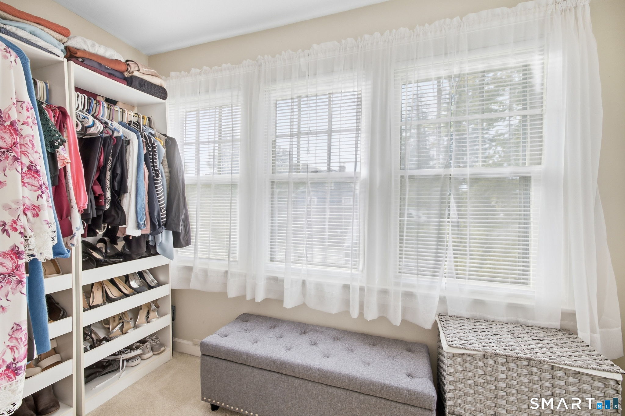 Walk-in closet with clothes and shoe shelves, a gray bench, and bright curtained windows.