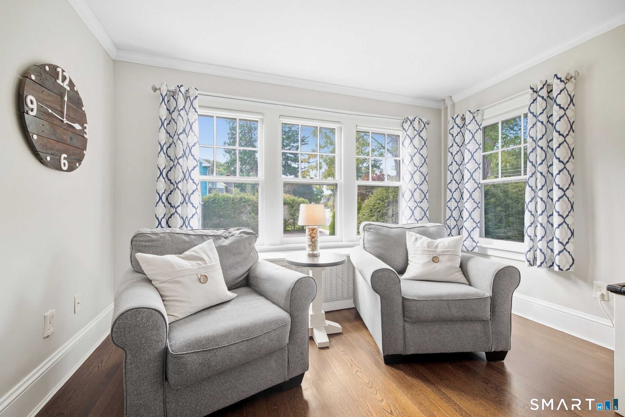 Furnished living room with two gray armchairs, patterned curtains, and large windows overlooking greenery.