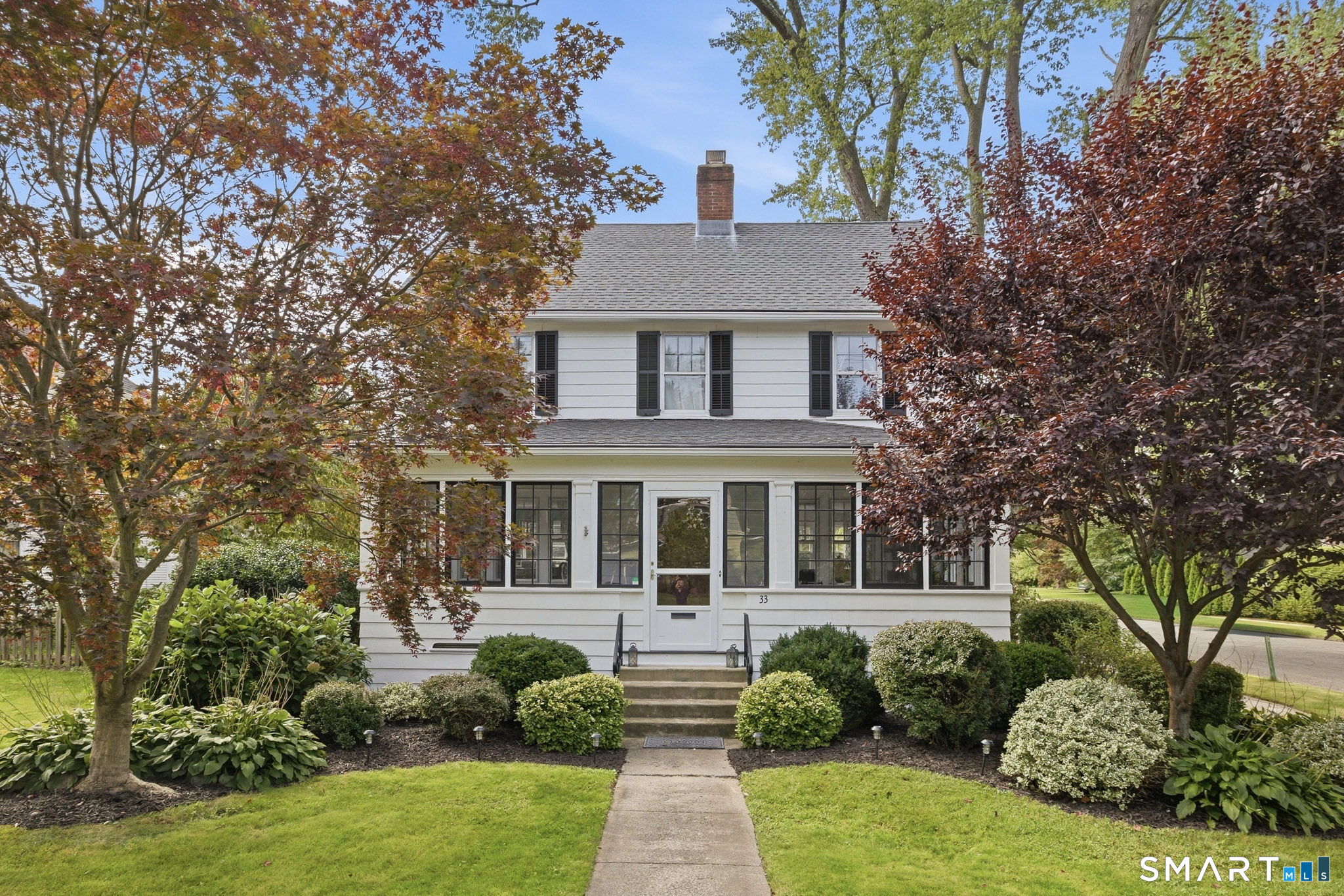 White two-story house with black shutters, a centered entrance, and autumn trees framing the front yard.
