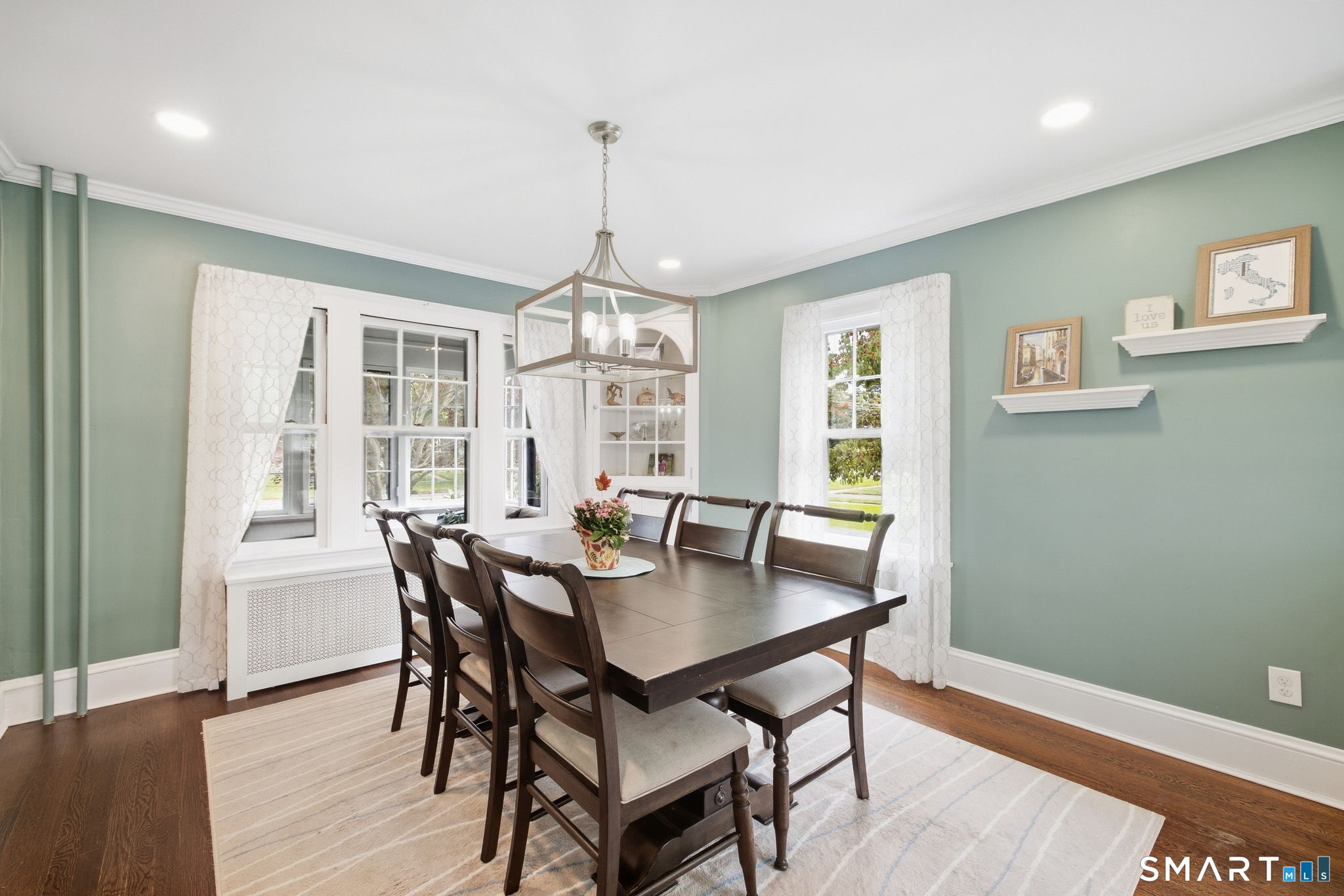 Dining room with a dark wood table, sage-green walls, large windows, and a geometric pendant light.