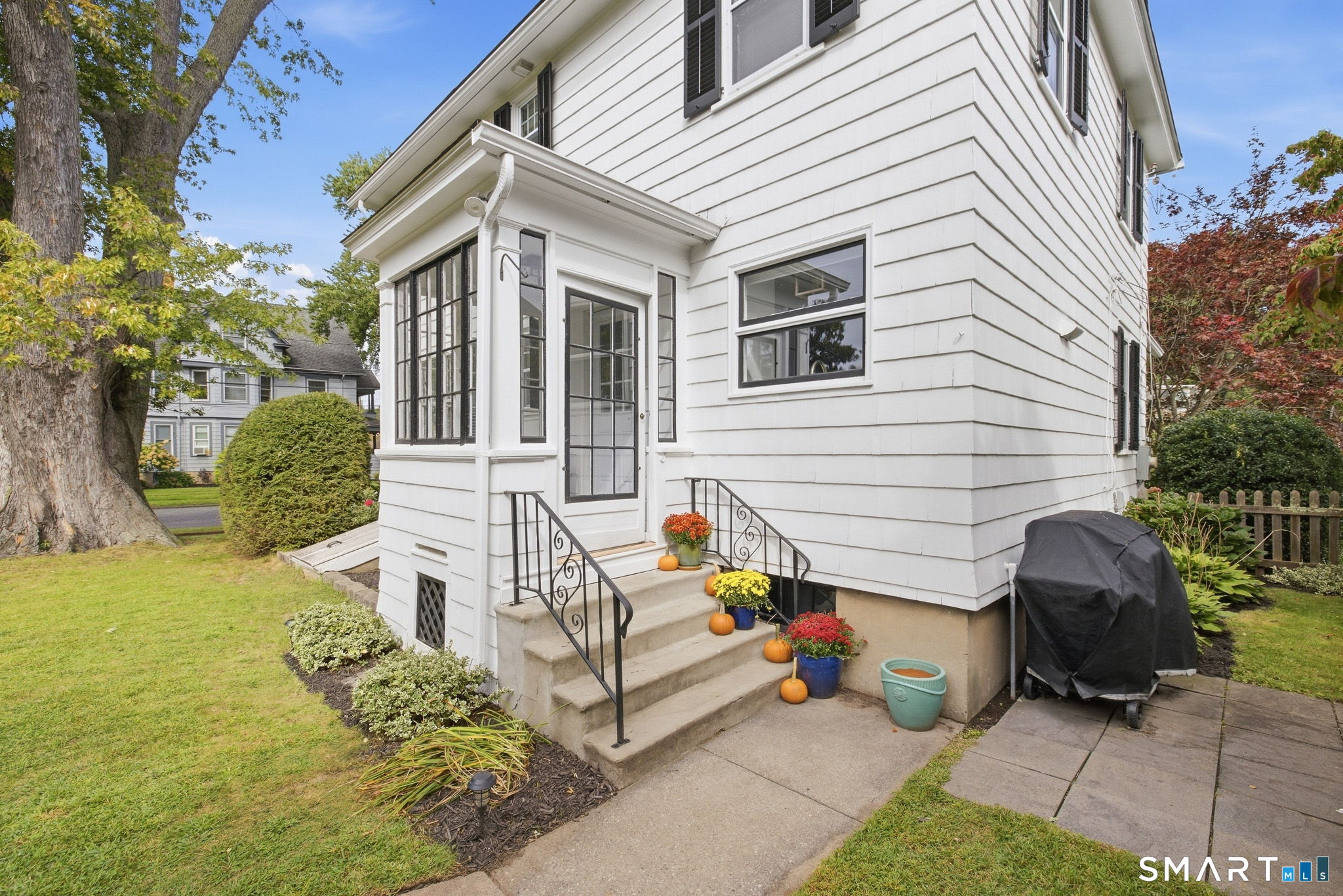 White two-story house with a glass-enclosed entry and flowerpots and pumpkins lining the steps.