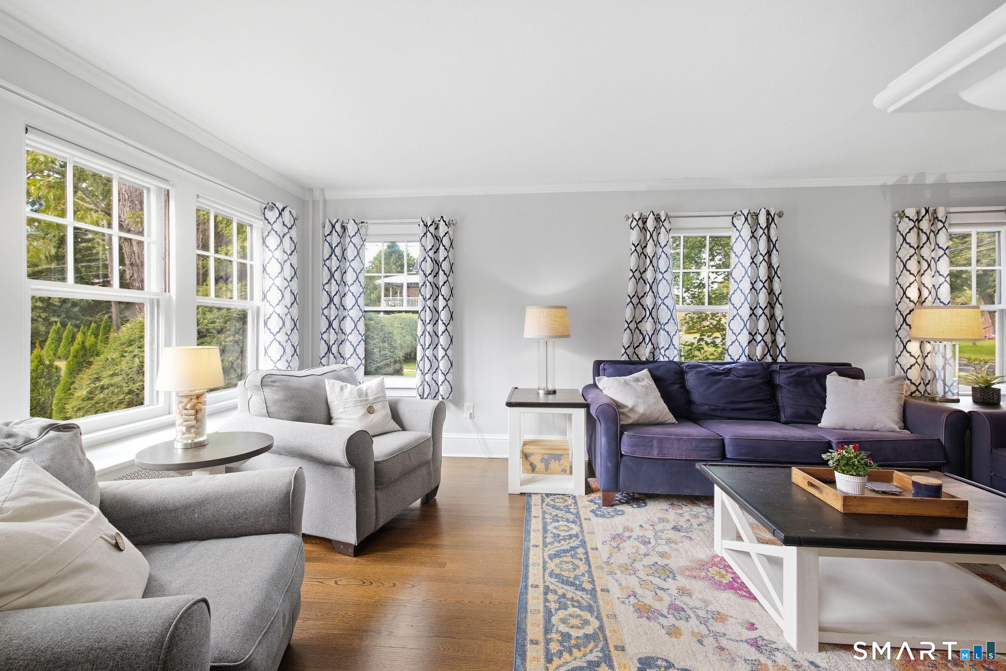 Living room with large windows, gray and purple sofas, patterned curtains, and hardwood floors.