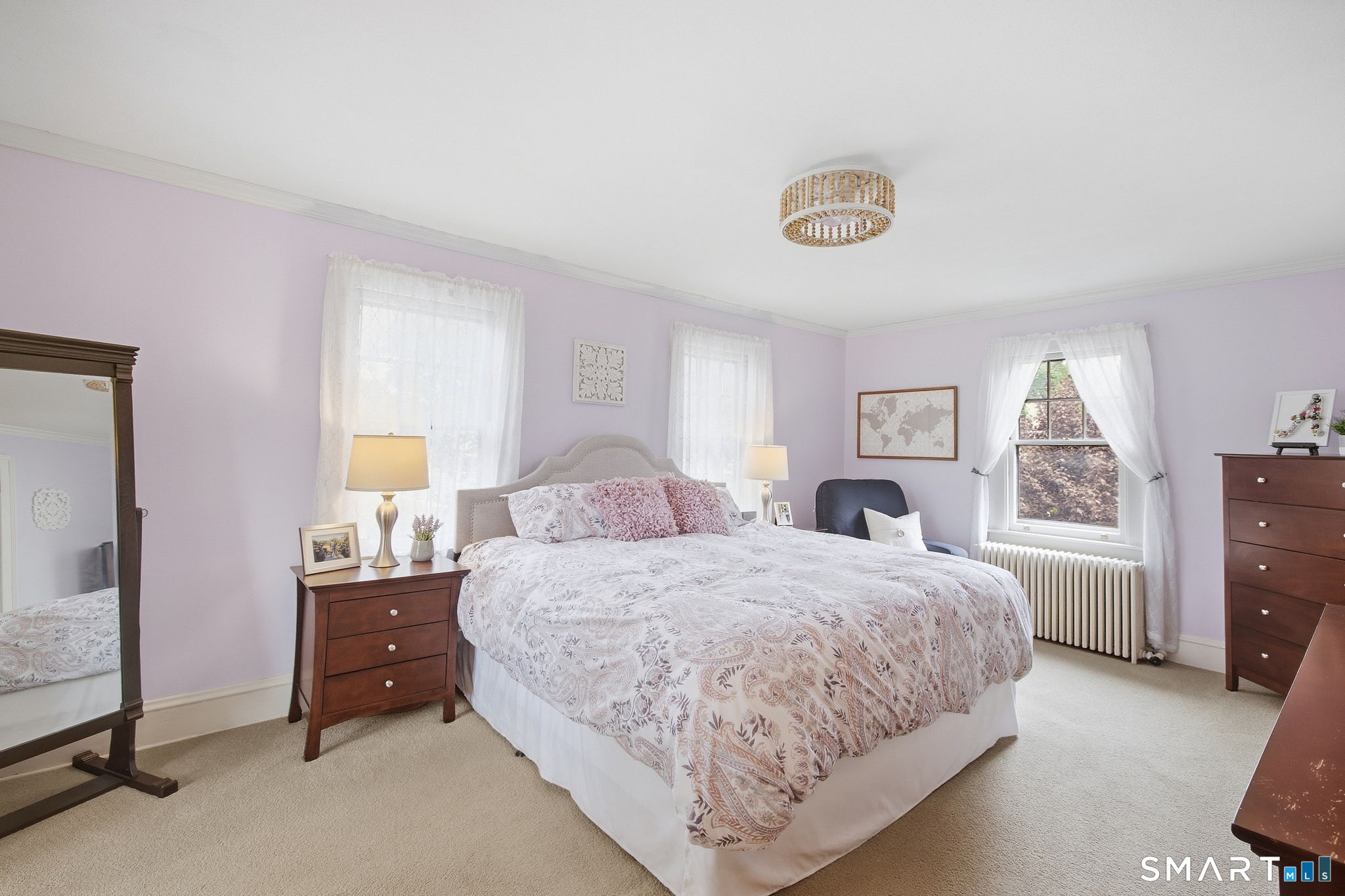 Bedroom with floral bedding, lavender walls, dark wood furniture, and three curtained windows.