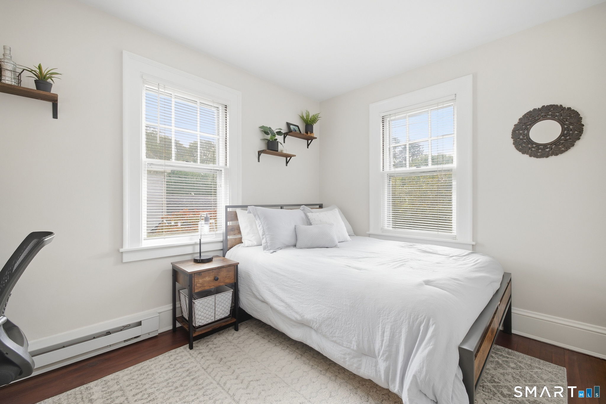 Bright bedroom with a white bed between two large windows and wooden bedside furniture.