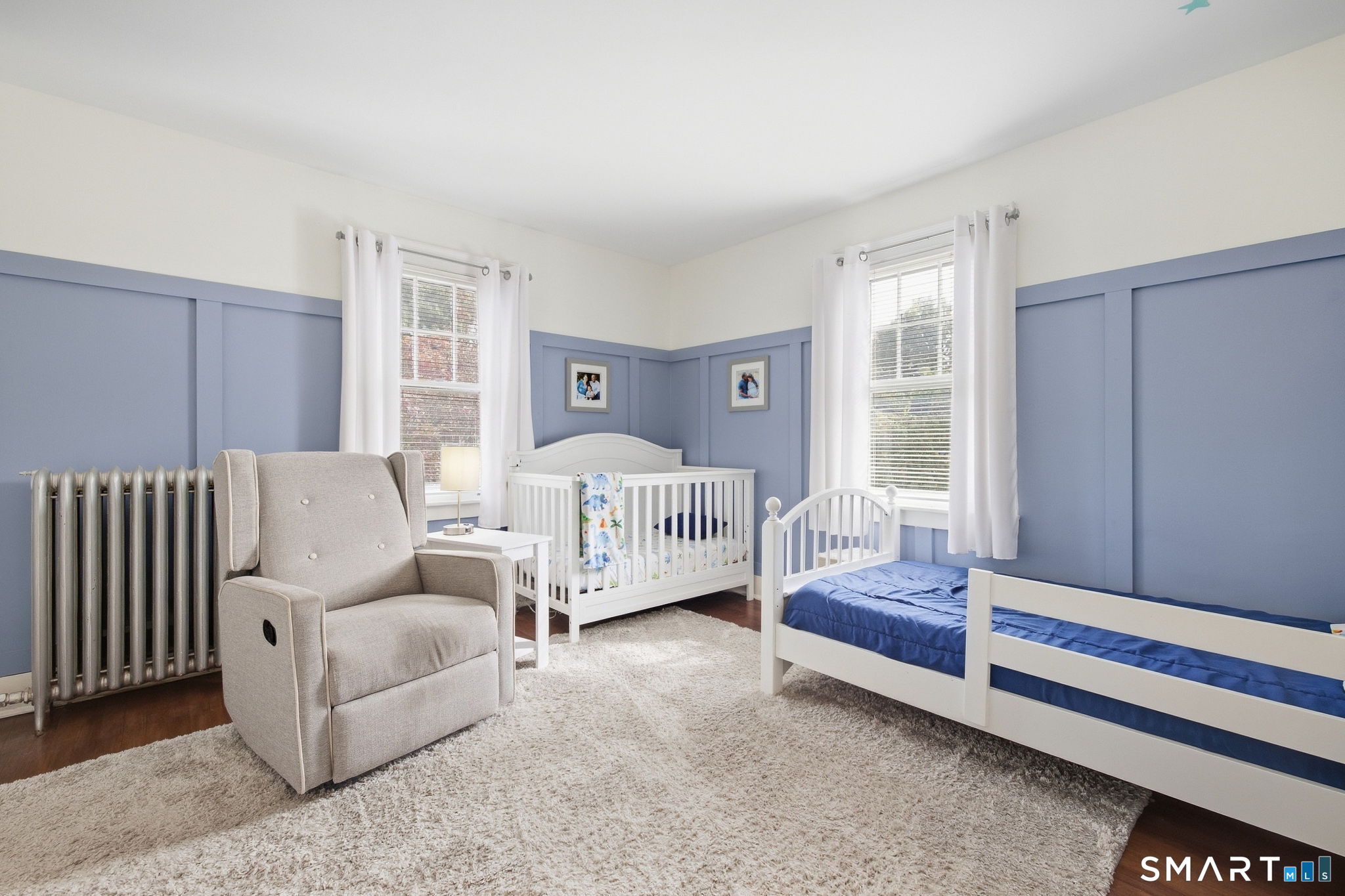 Nursery with blue paneled walls, a white crib and twin bed, and a beige rocking chair.